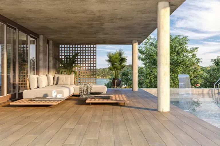 Un patio moderno con suelo de madera clara, sofá de exterior, mesas de madera y una piscina con vistas a un paisaje verde.