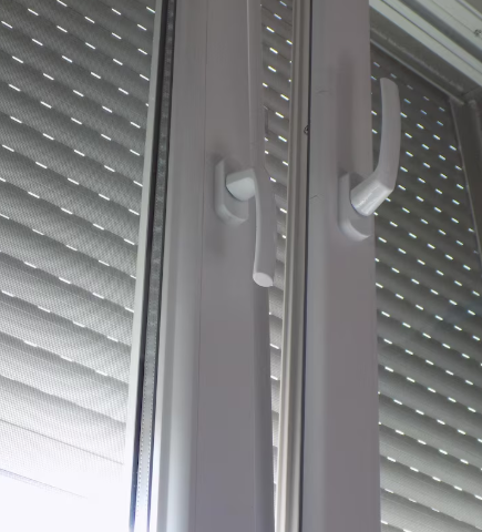 Marco de ventana blanco con dos manijas, sobre una cubierta de ventana con listones de color gris.