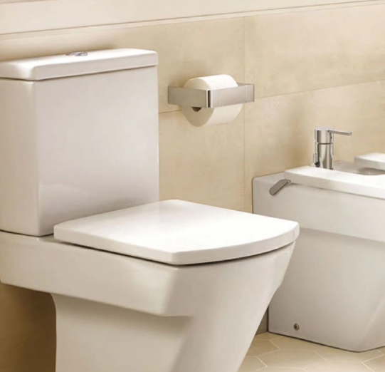 Inodoro blanco con tapa cerrada, un rollo de papel higiénico y un bidé en un baño.