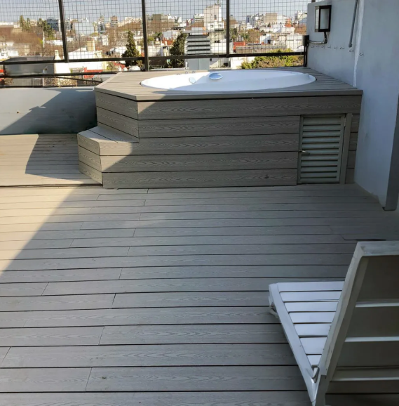 Un jacuzzi blanco sobre una terraza de material compuesto gris con un moderno sillón y vistas a la ciudad desde una terraza vallada.