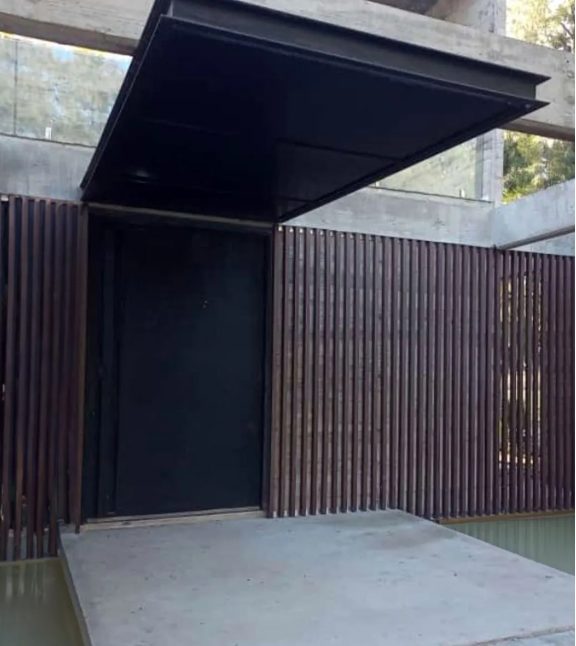 Una puerta moderna oscura con listones de madera verticales, un porche de hormigón y un alero de metal negro debajo de una estructura de hormigón.
