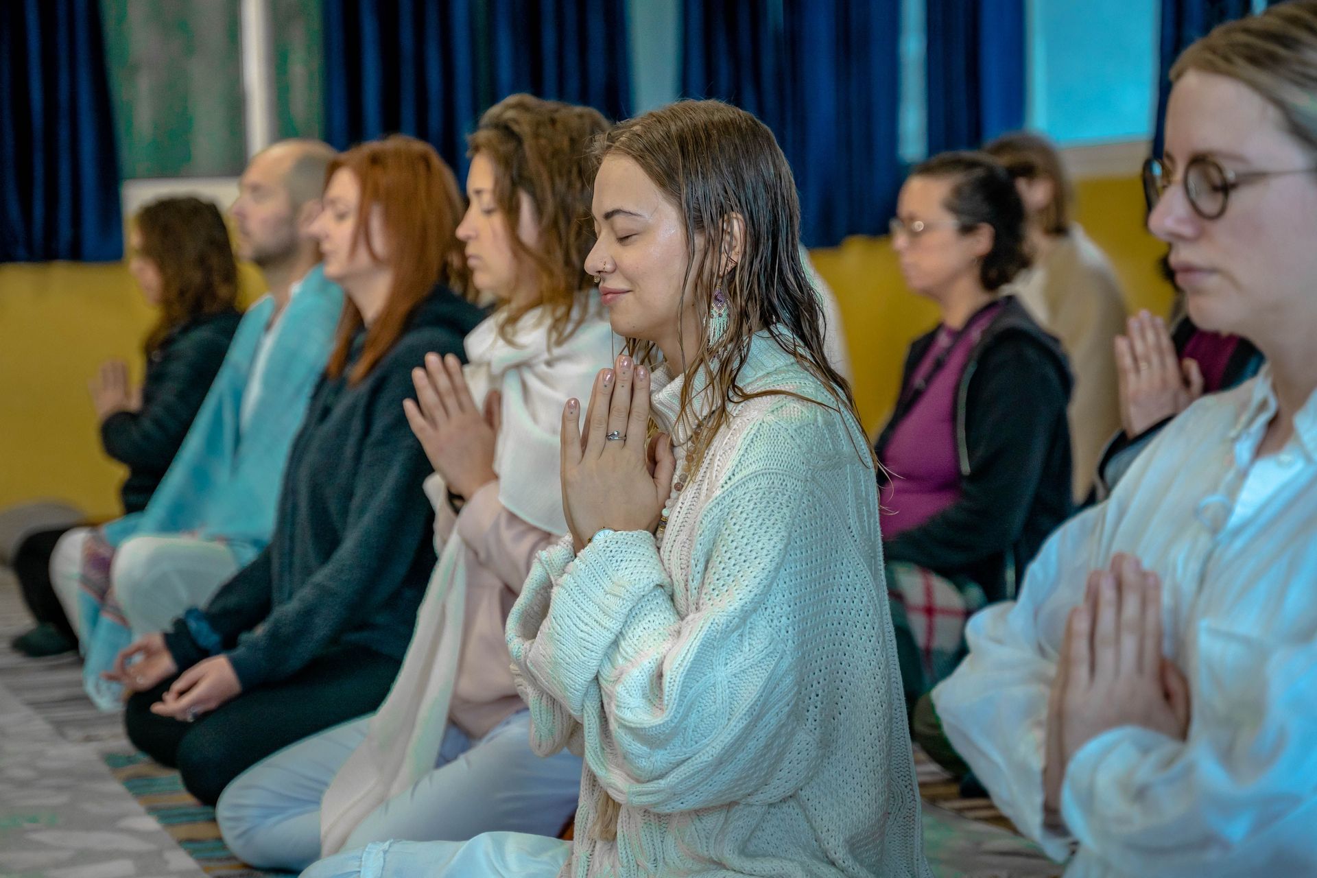 People seated in meditation, hands pressed together, in a calm indoor setting