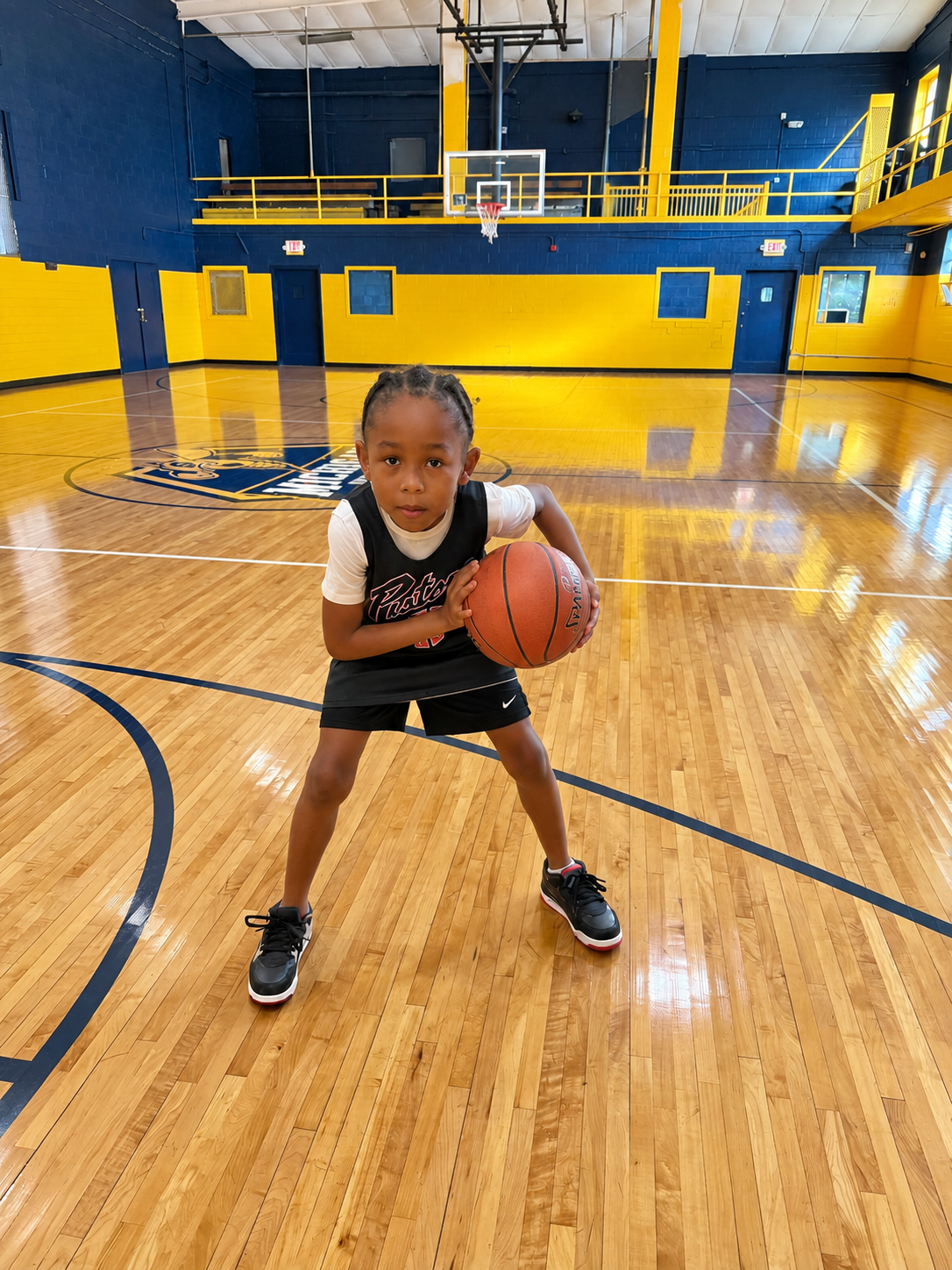 Child dribbling a basketball in a blue-and-yellow gym, crouched low on the court.