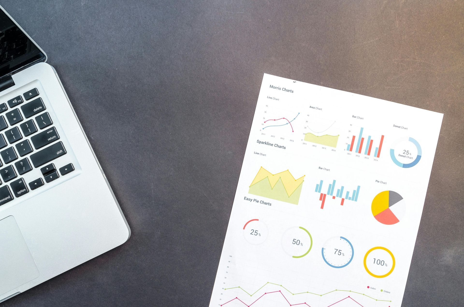 Laptop and report with colorful charts on a dark gray surface, suggesting business analysis.