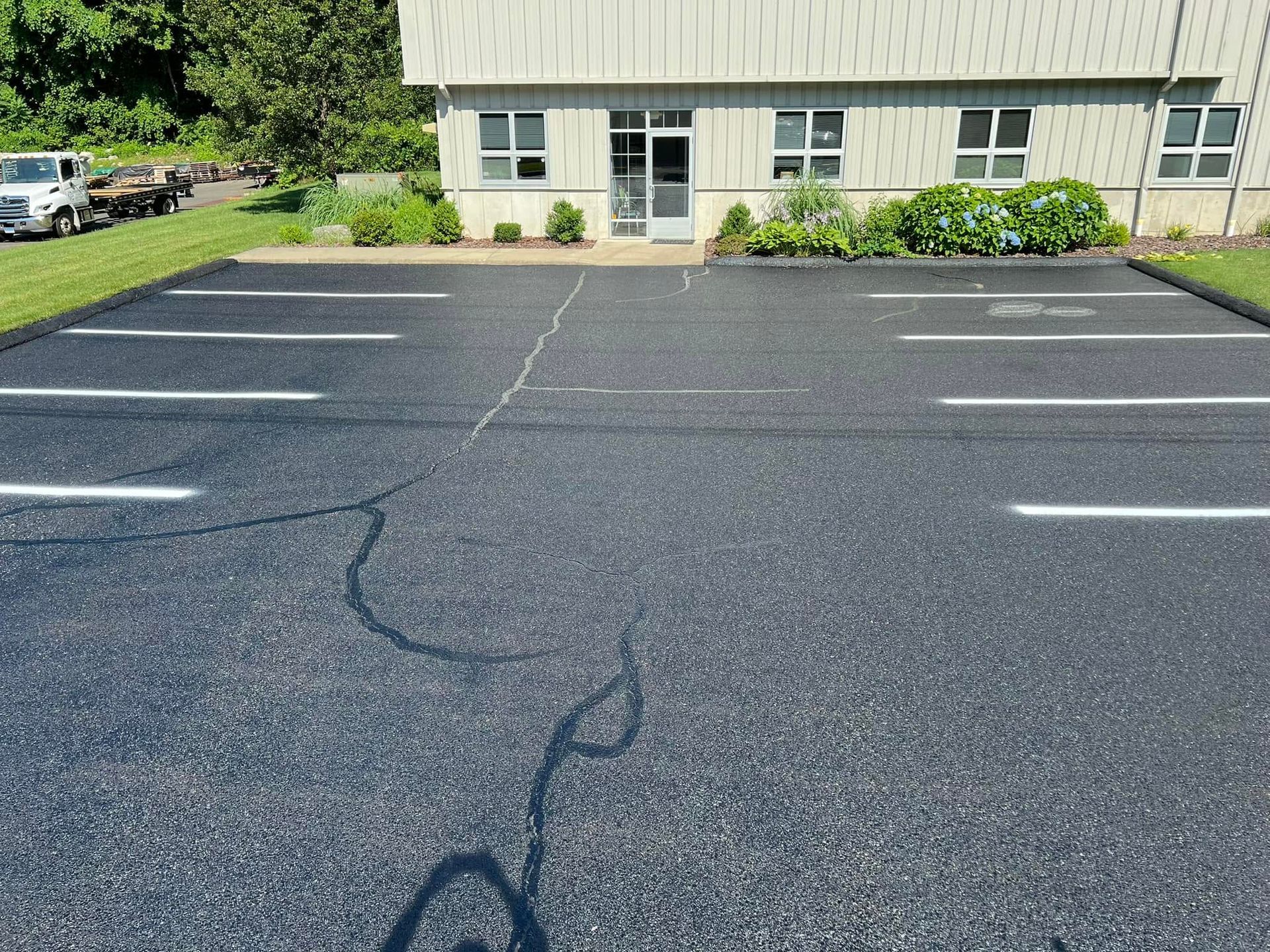 Empty asphalt parking lot with cracked pavement in front of a light-colored building and greenery