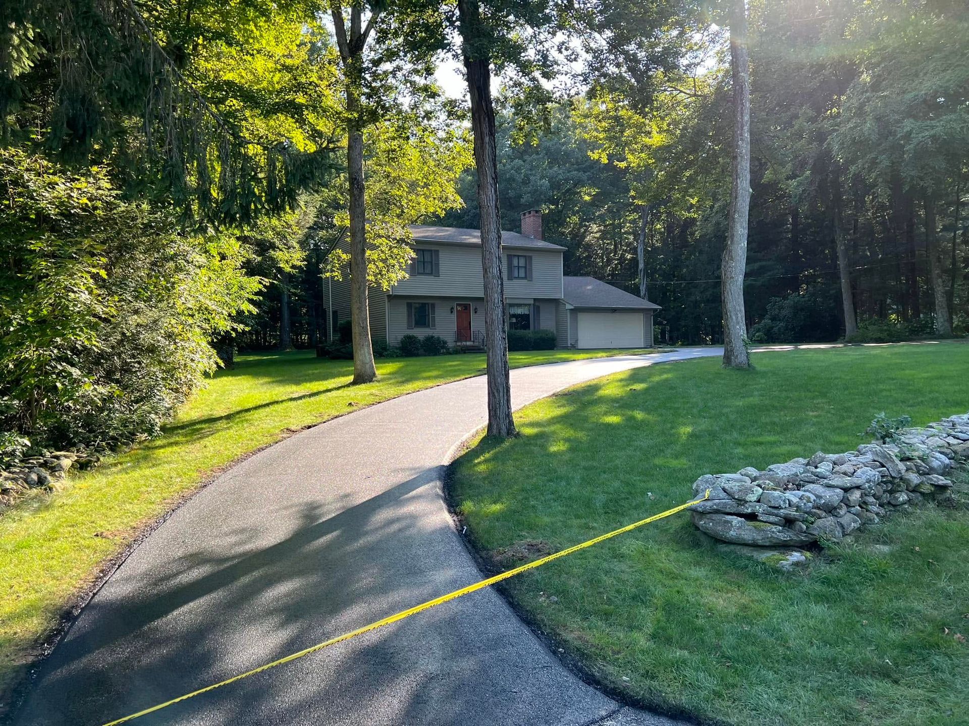 Curved driveway leading to a house in a wooded yard, with green grass and trees in sunlight