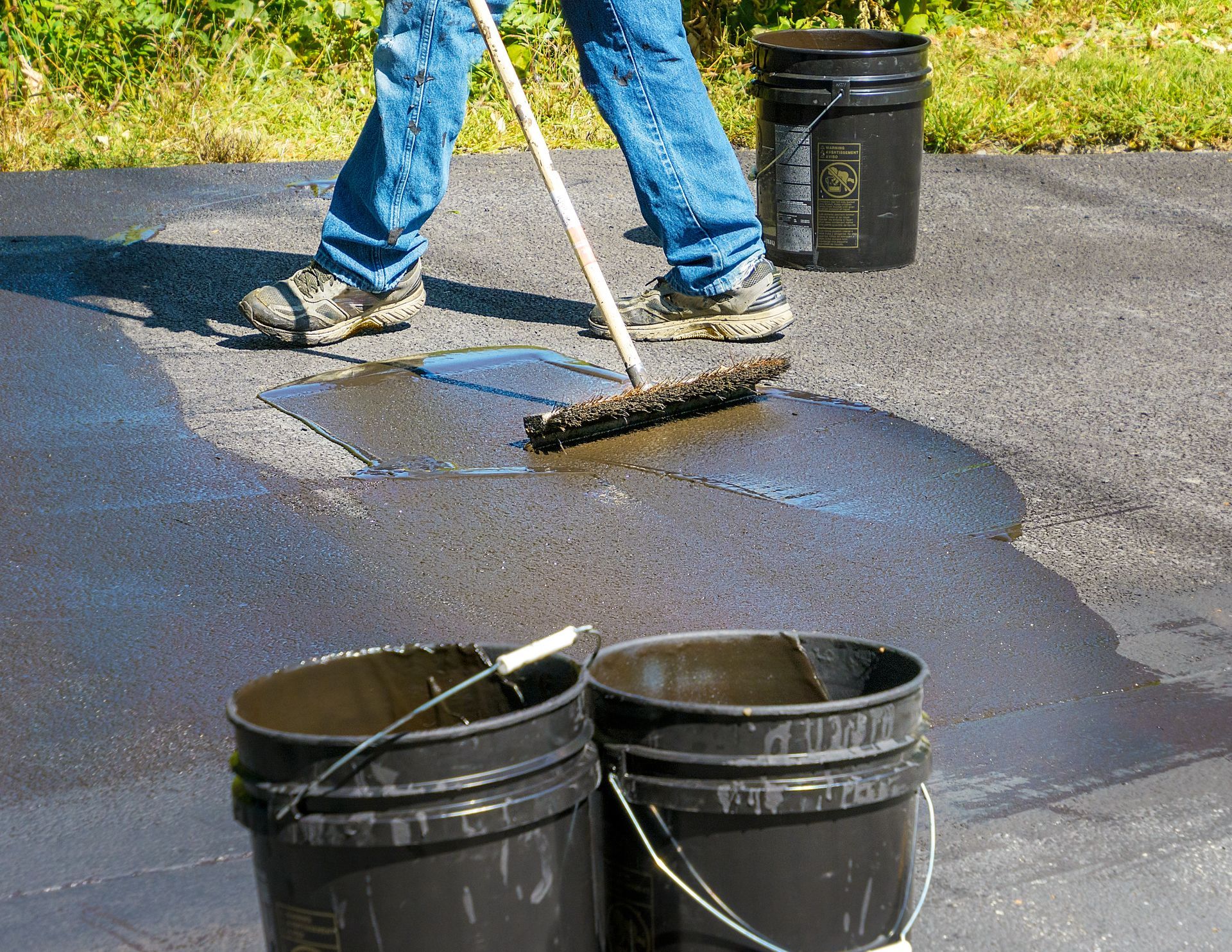 Person spreading black sealant on pavement with a broom, with buckets nearby.