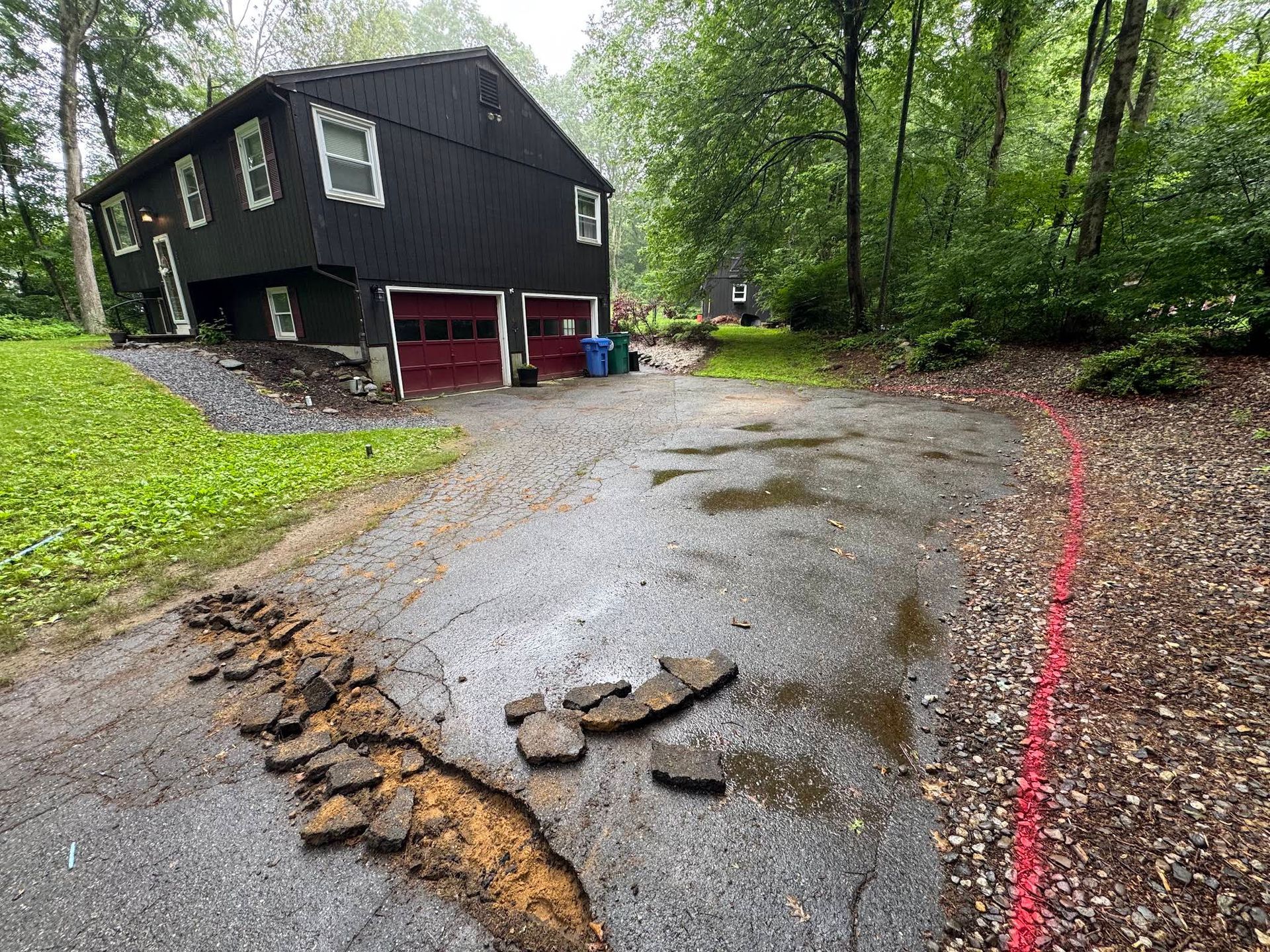 Black house beside a wet, cracked driveway in a wooded area after erosion or flood damage