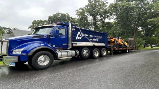 Blue tow truck hauling heavy equipment on a wet road in a park-like setting