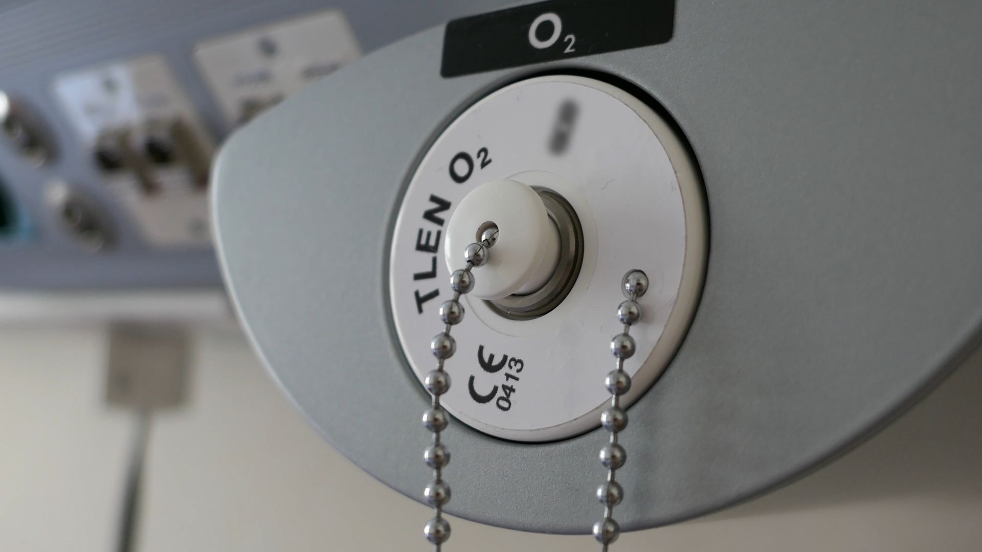 Oxygen gas outlet labeled “O2” with connected tubing and chain in a medical setting