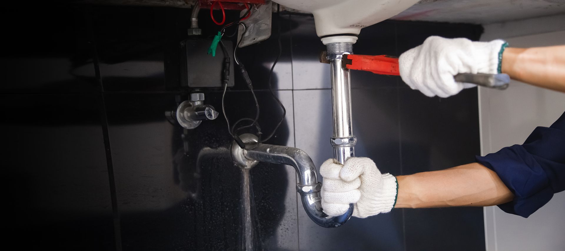 Gloved hands tightening a pipe under a sink with a wrench in a dark utility area
