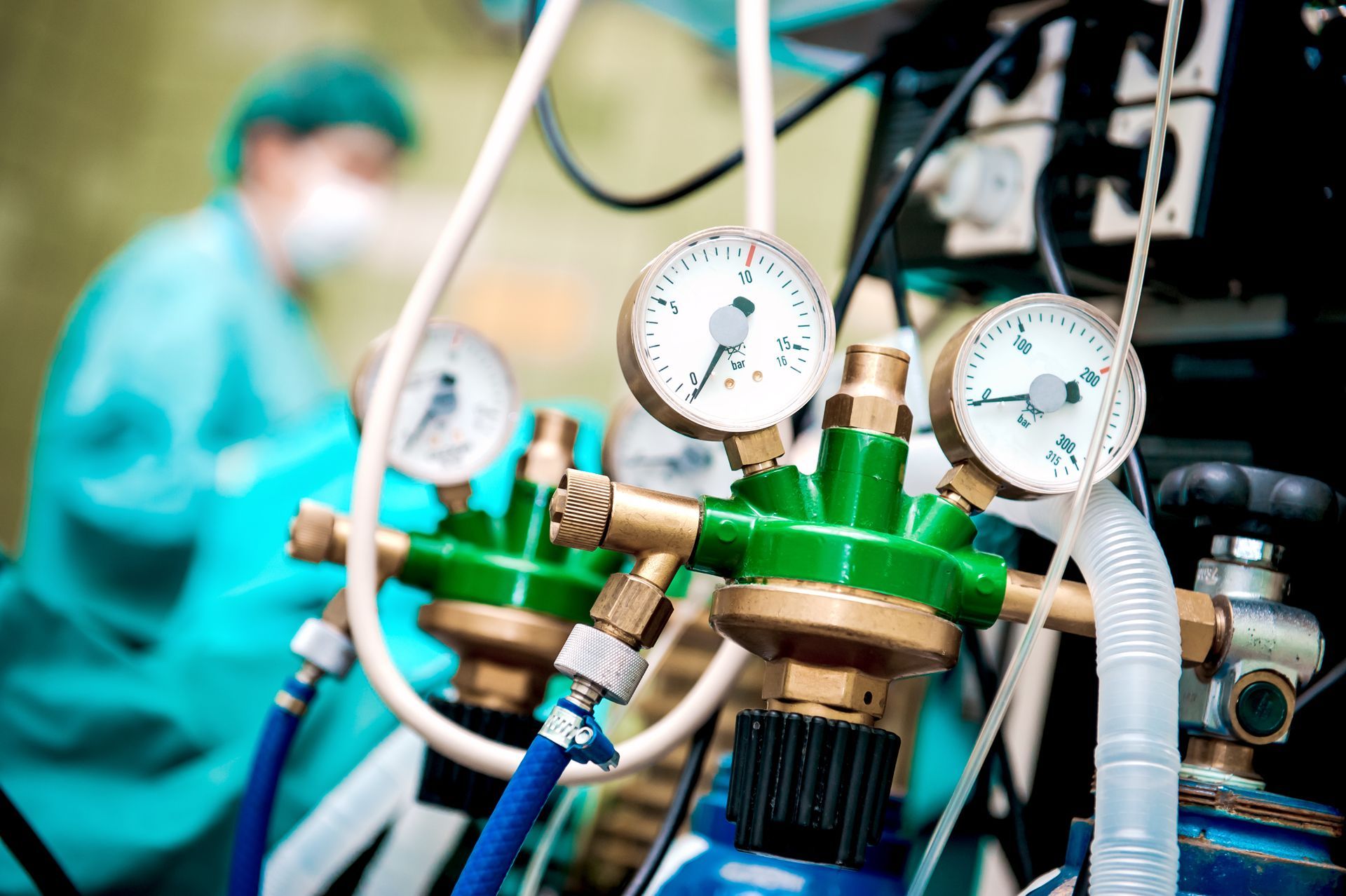 Medical gas regulators with gauges and blue tubing in a clinical setting, technician blurred in background
