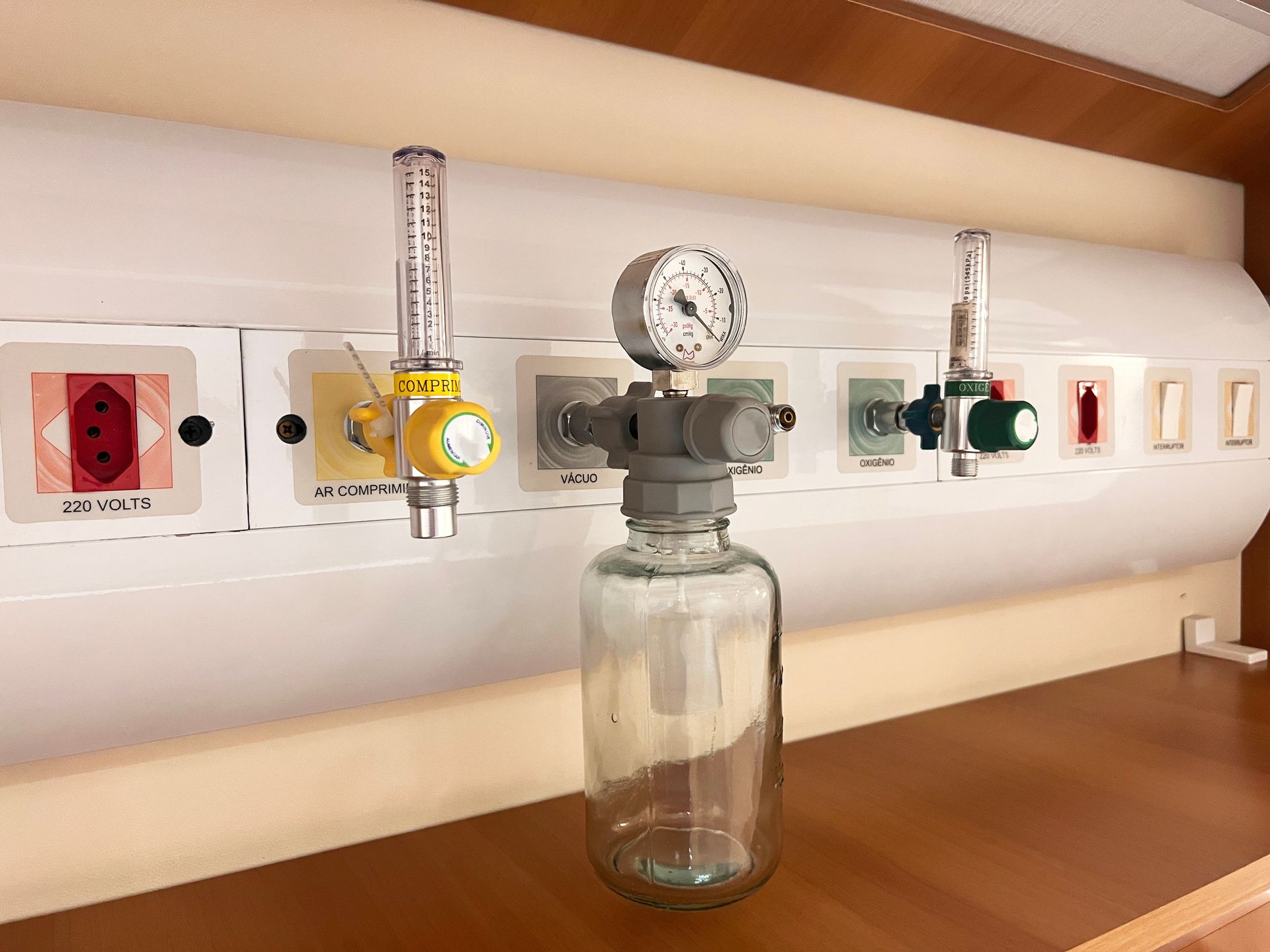 Close-up of a wall-mounted lab gas outlet with pressure gauge and glass bottle beneath, in a wooden case
