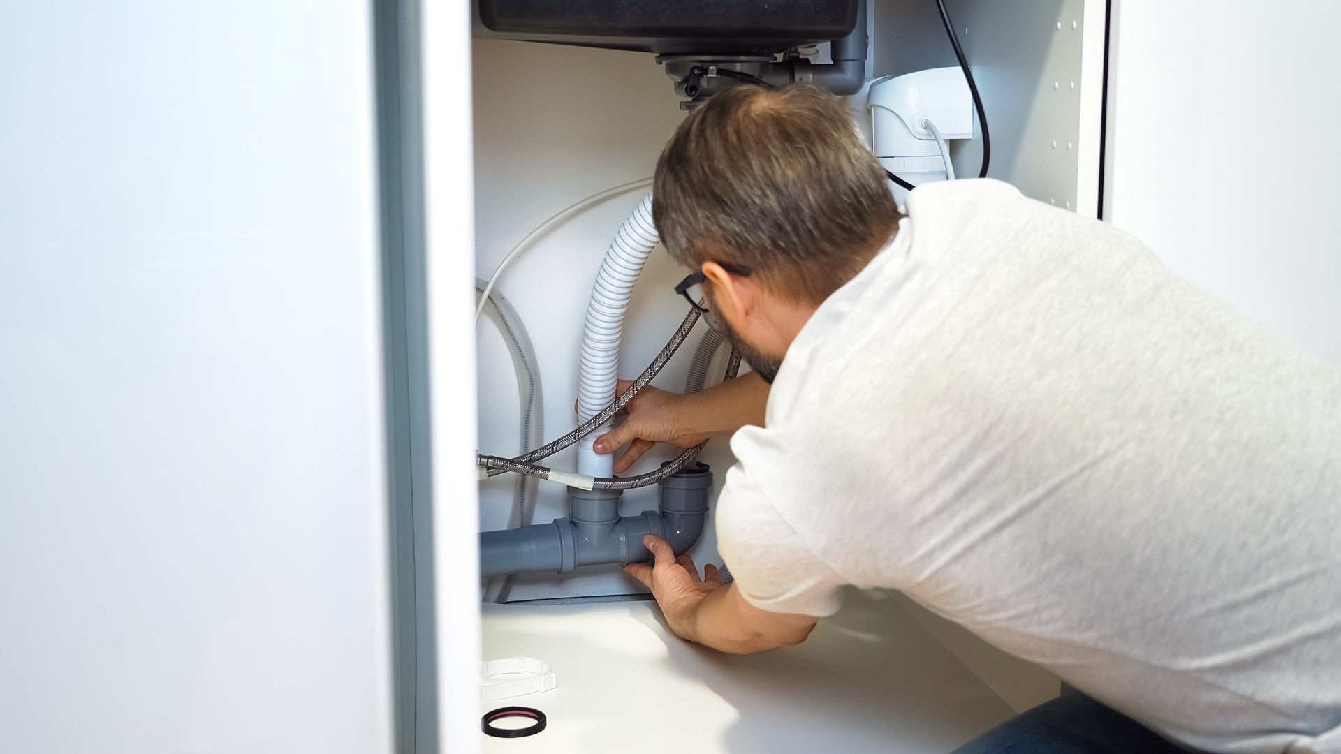 Person working under a sink with a tool, adjusting plumbing inside a cabinet.
