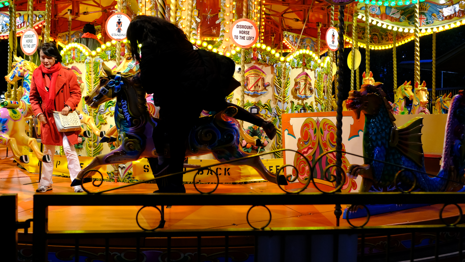 Woman in red coat beside a carousel with fantasy creatures lit at night.