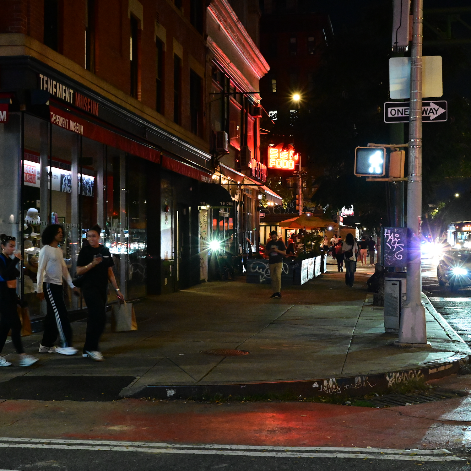Nighttime street scene. People walk along a lit sidewalk near storefronts and crosswalk. A car passes by.