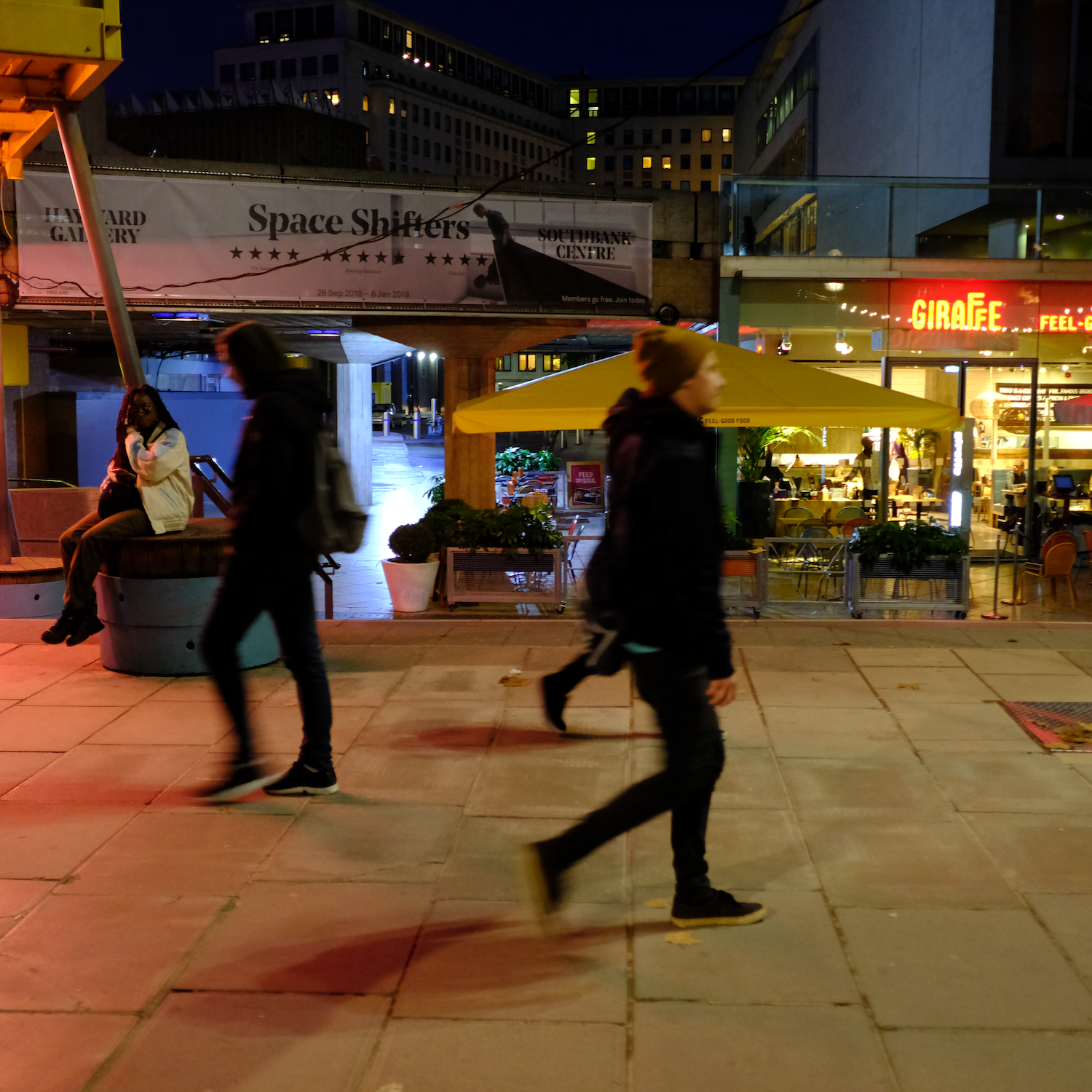 People walking on a city sidewalk at night, with illuminated shops and buildings in the background.