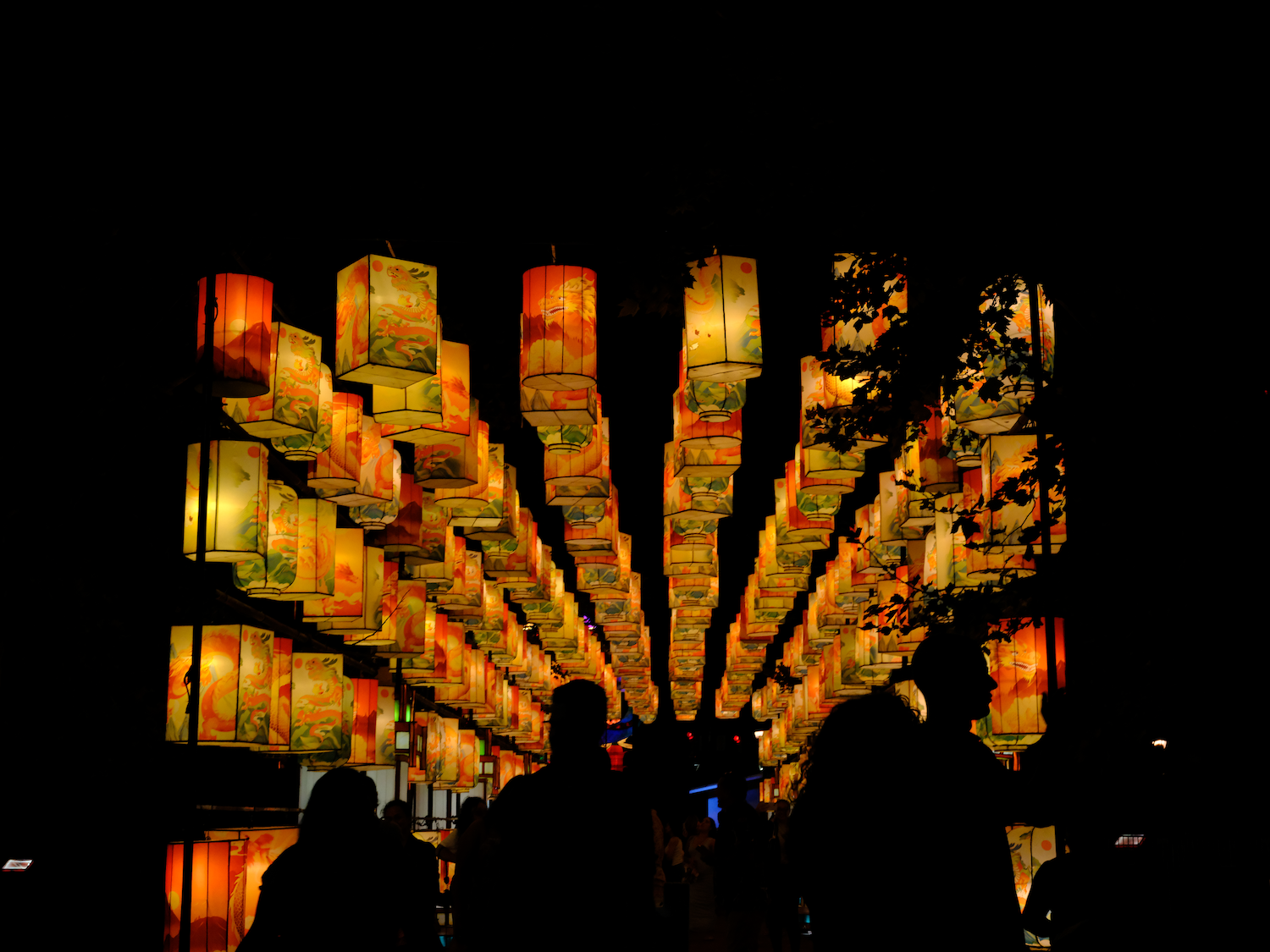 Rows of illuminated orange and yellow paper lanterns hanging outdoors at night. Silhouetted figures stand below.