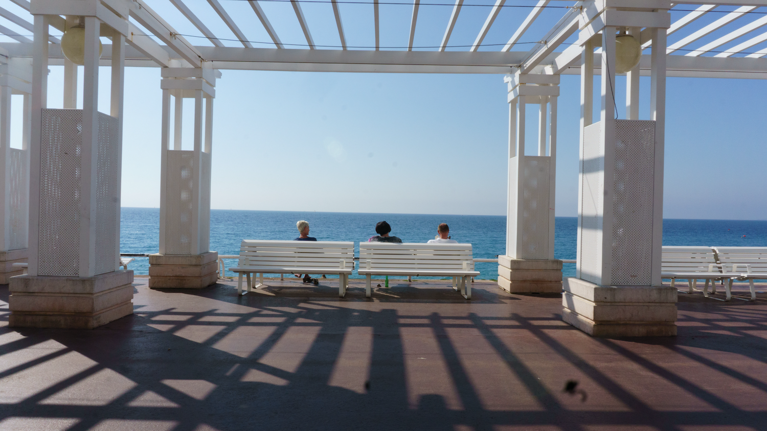 White pergola framing three people seated on a bench, looking out at the sea. Blue sky.