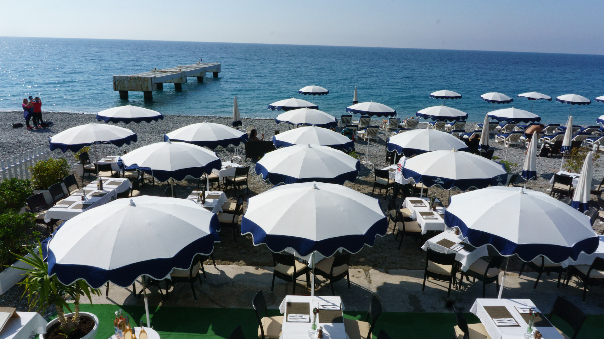 Beachfront restaurant with white and navy umbrellas over tables, near a pebbled shore and ocean.