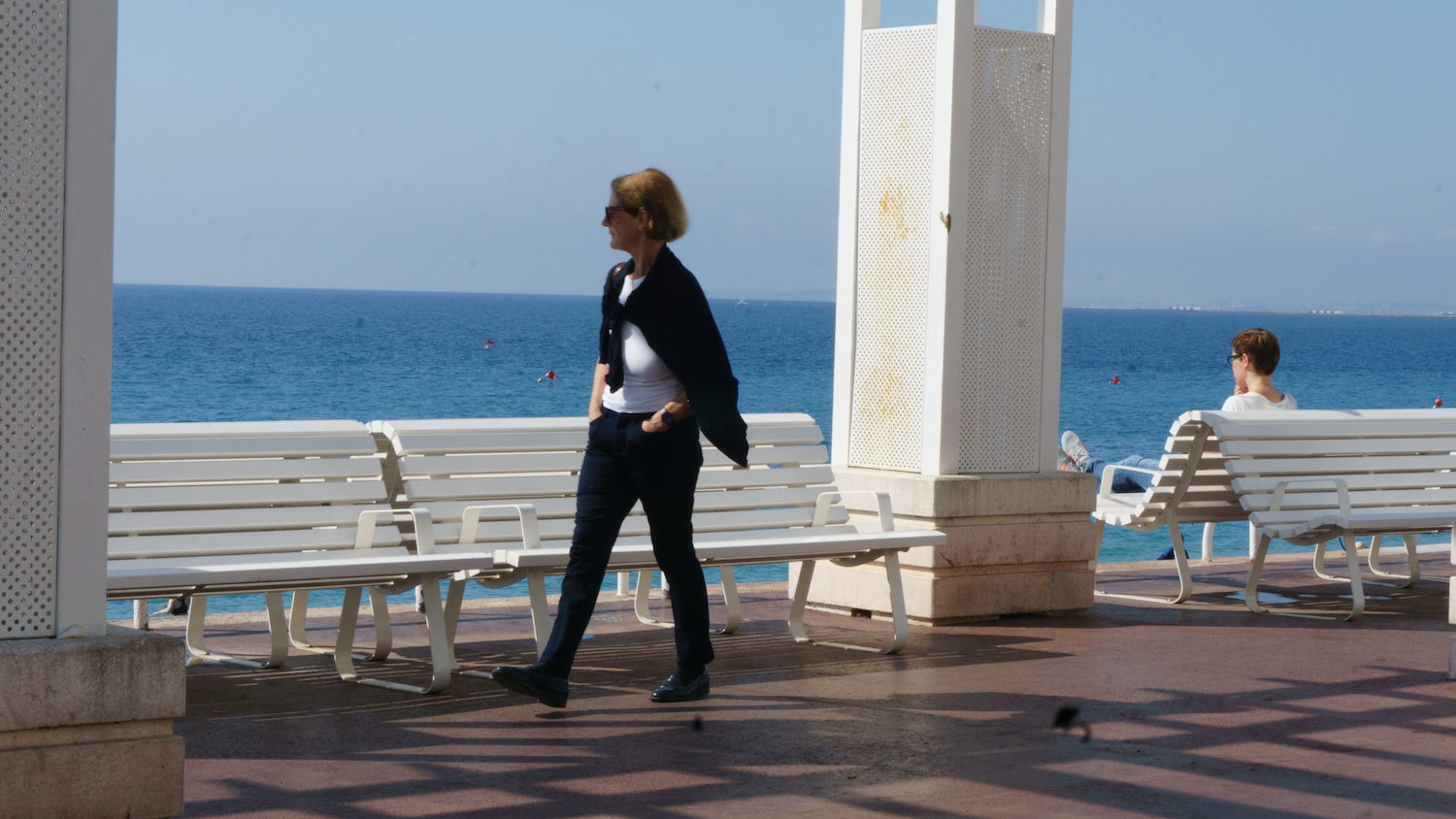 Woman walking on a seaside promenade; blue sea and sky in the background.