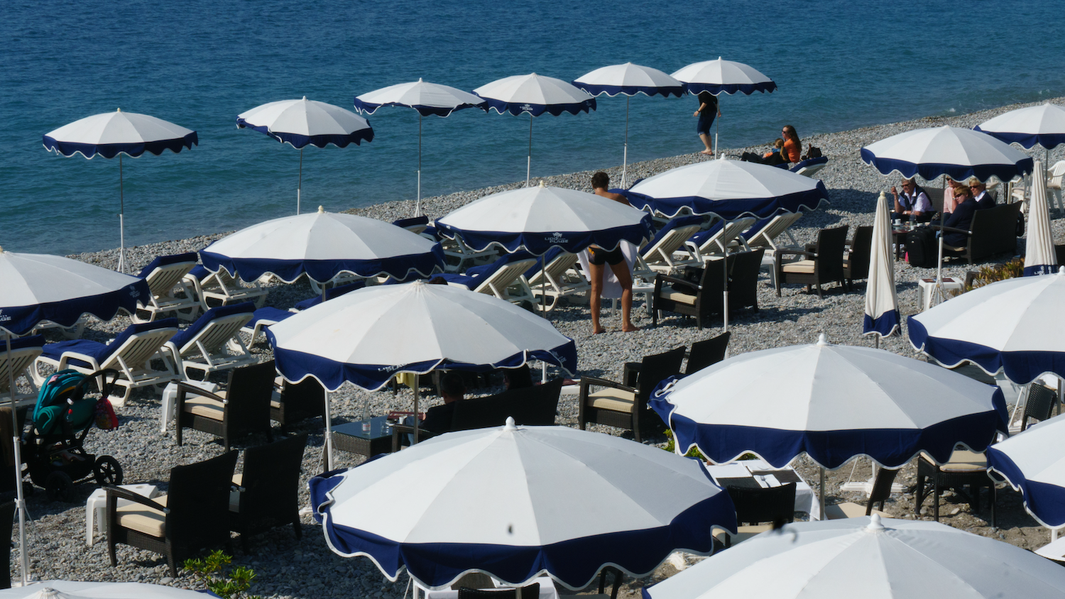 Beach with white umbrellas, blue trim, lounge chairs, and people near the water's edge.