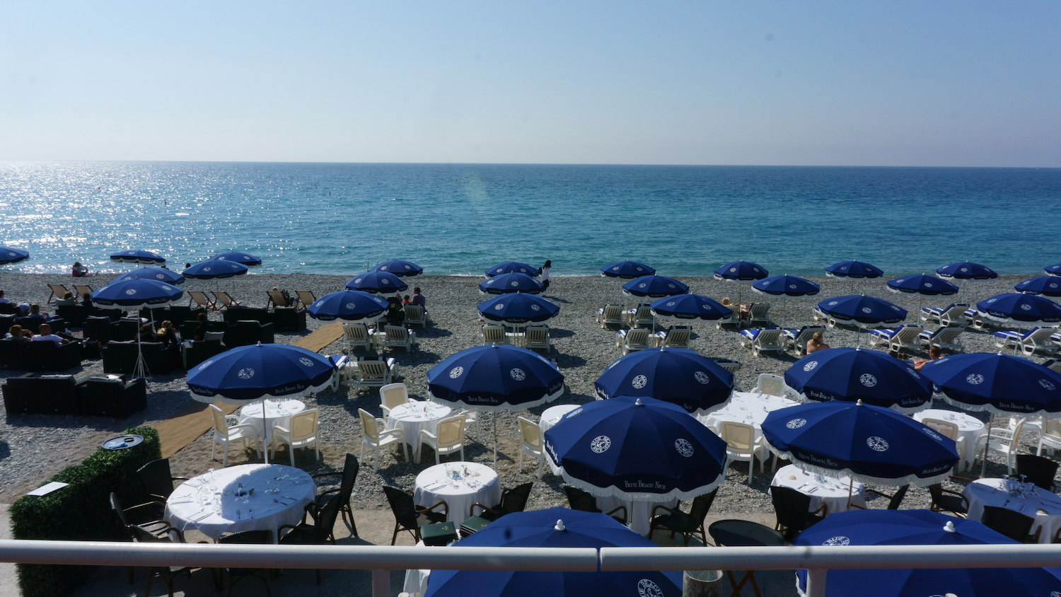 Beach umbrellas dot a shoreline with tables and chairs near the water. Blue ocean and sky.