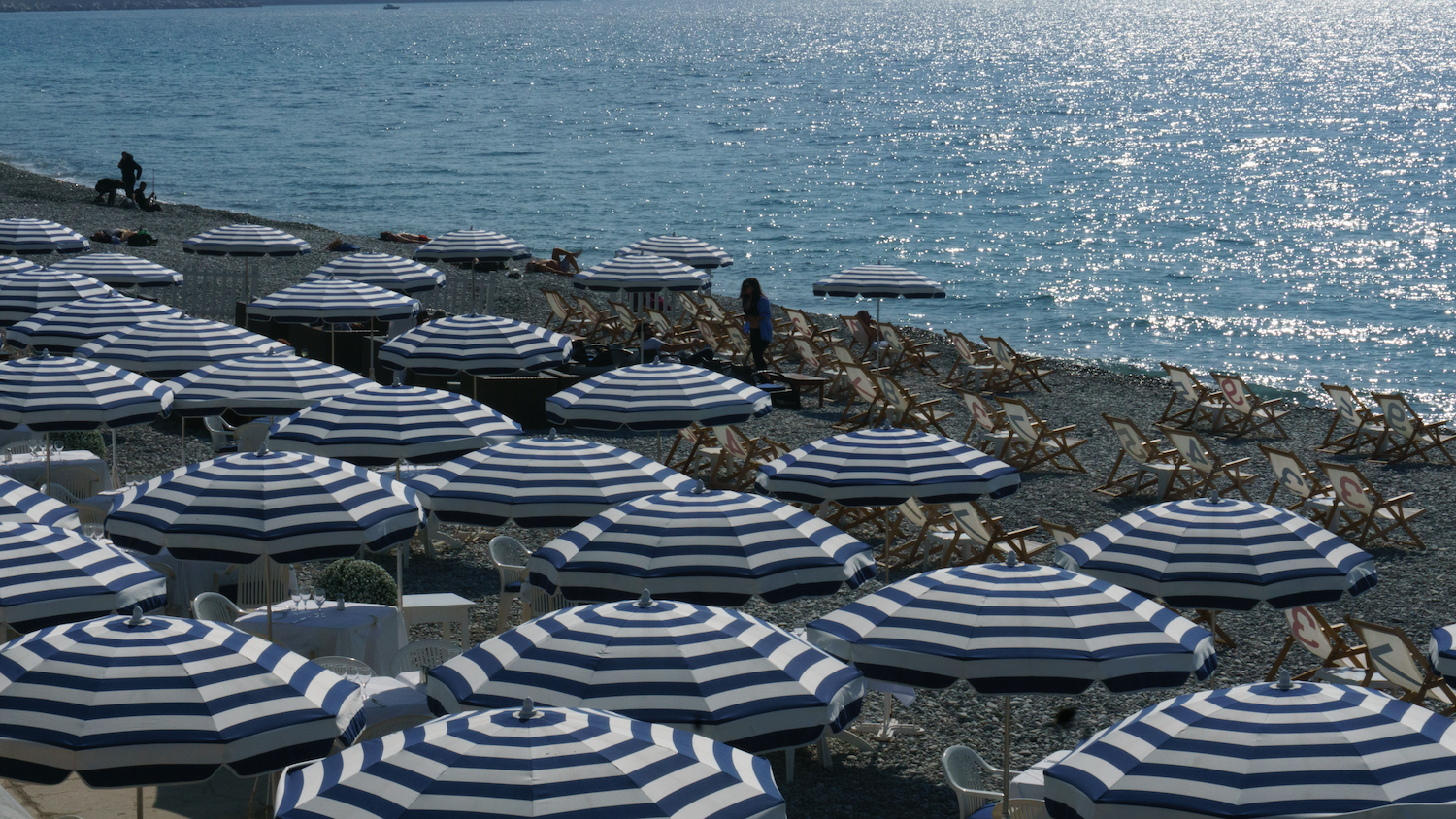 Rows of blue and white striped umbrellas on a rocky beach, sparkling ocean in the background.