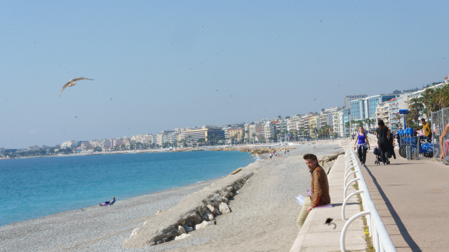 Seaside view with a beach, buildings, and a person sitting on a wall. Turquoise water and a clear sky.