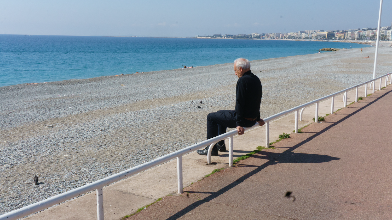 Man sitting on railing, looking out at a pebble beach and turquoise sea. City skyline in the background. Sunny day.