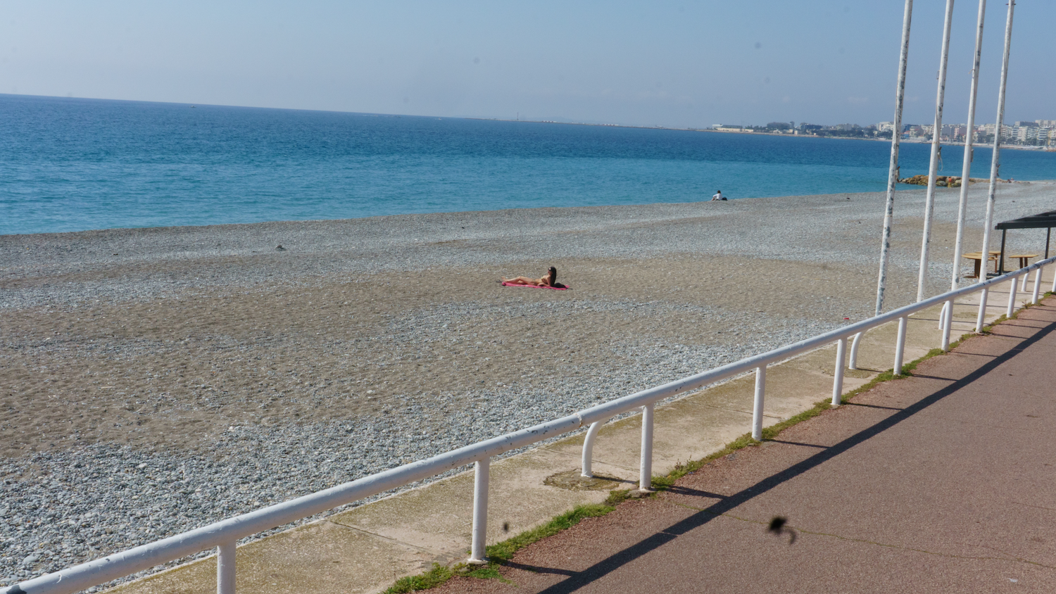Beach scene, turquoise water, pebble beach, person sunbathing, white railings and poles, blue sky.