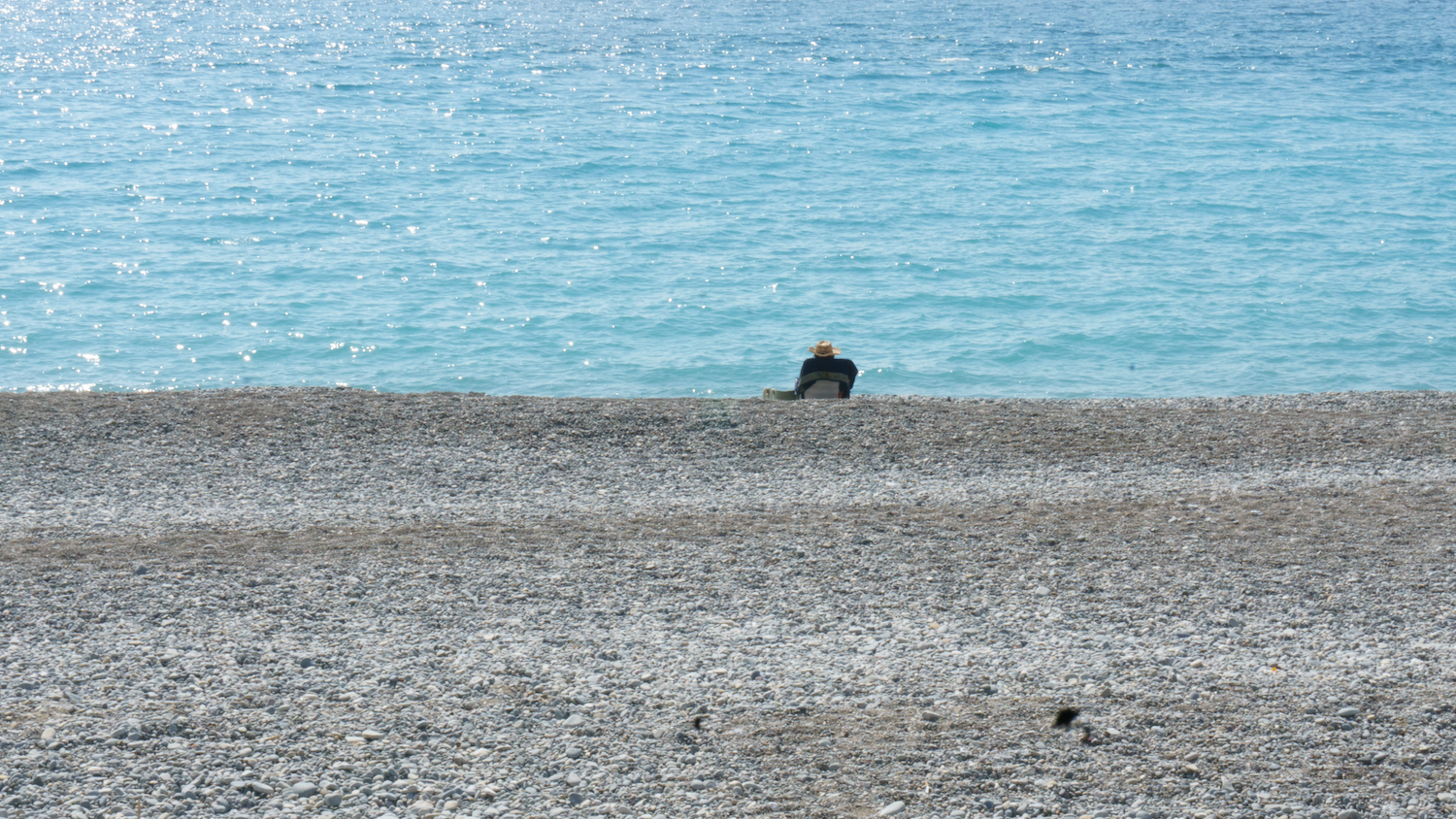 Person seated on a pebble beach, looking at a sparkling turquoise sea.