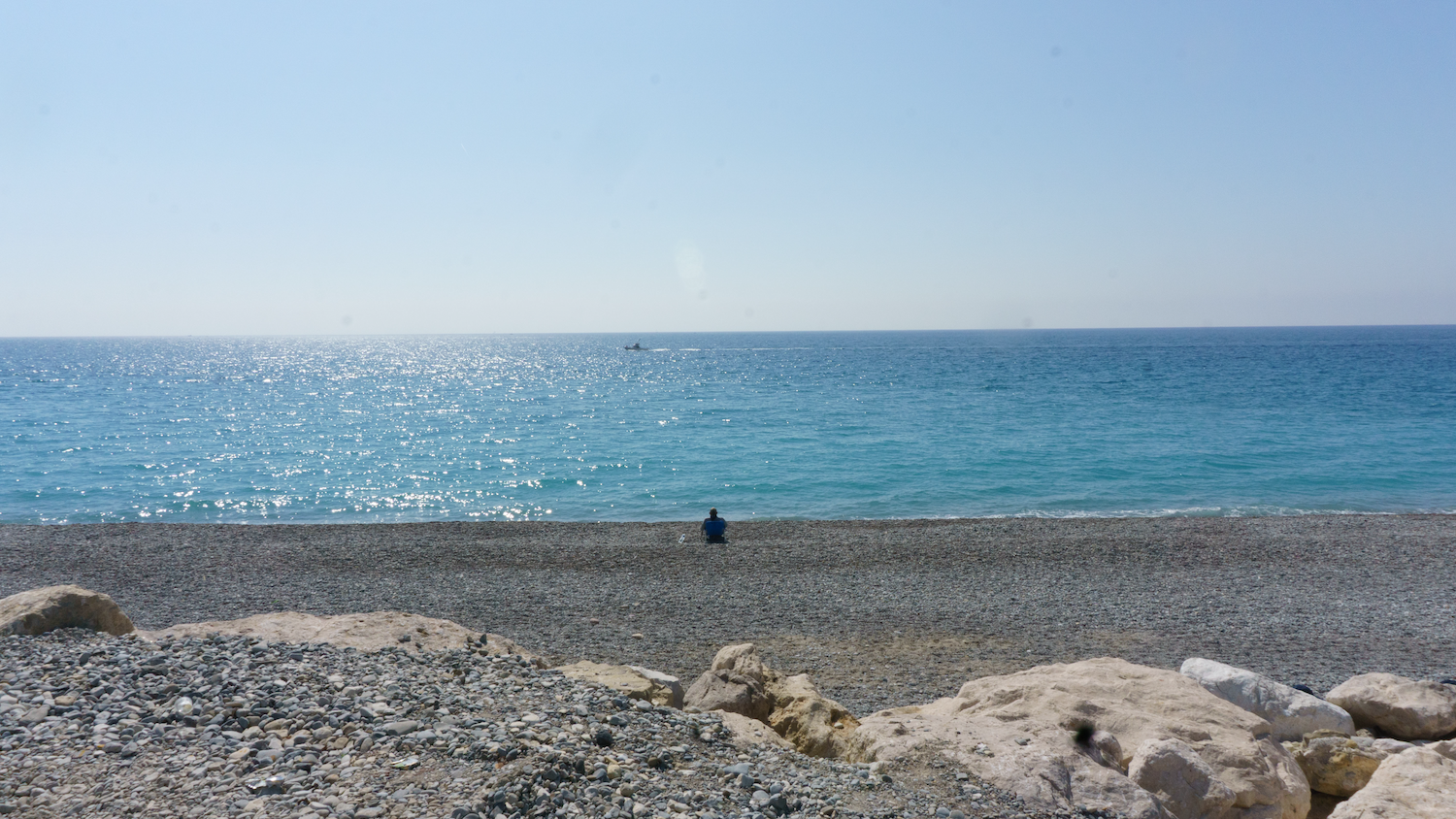 A person sits on a pebbled beach, facing the sparkling blue sea under a bright sky.