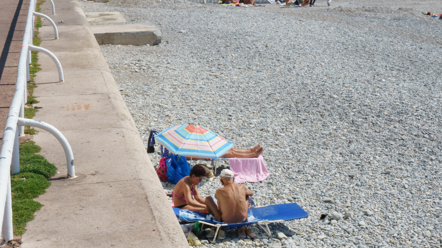 People relaxing on a pebble beach, next to a concrete wall with a railing. Blue beach umbrella, sunny day.