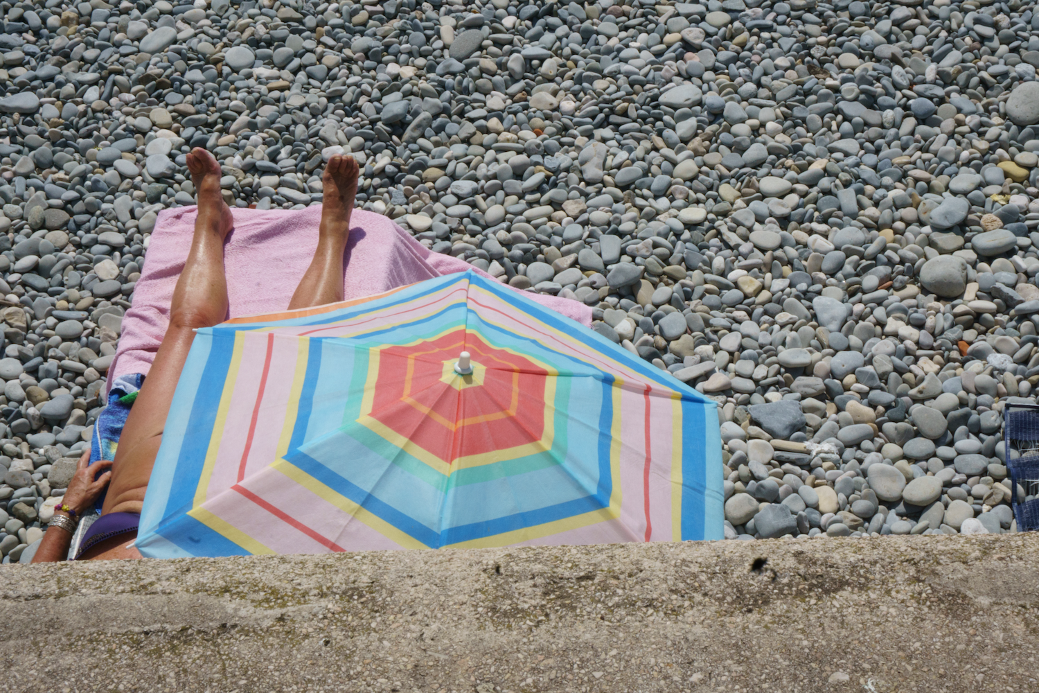 Person lying on a pebble beach under a striped umbrella.
