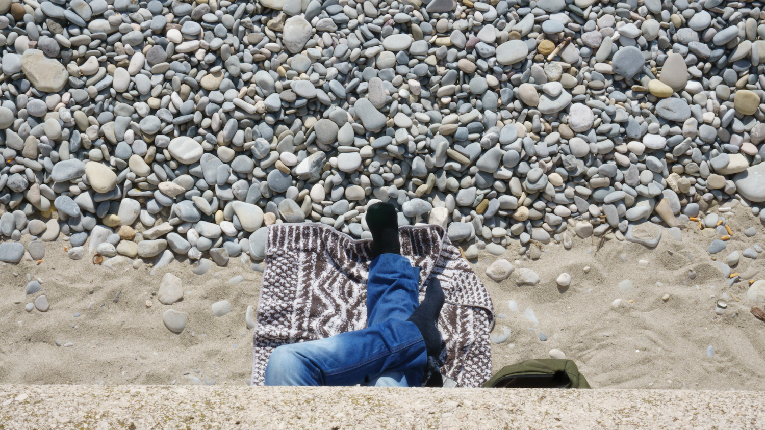 Person's legs in blue jeans and black socks on a patterned blanket at the edge of sand and pebbles.