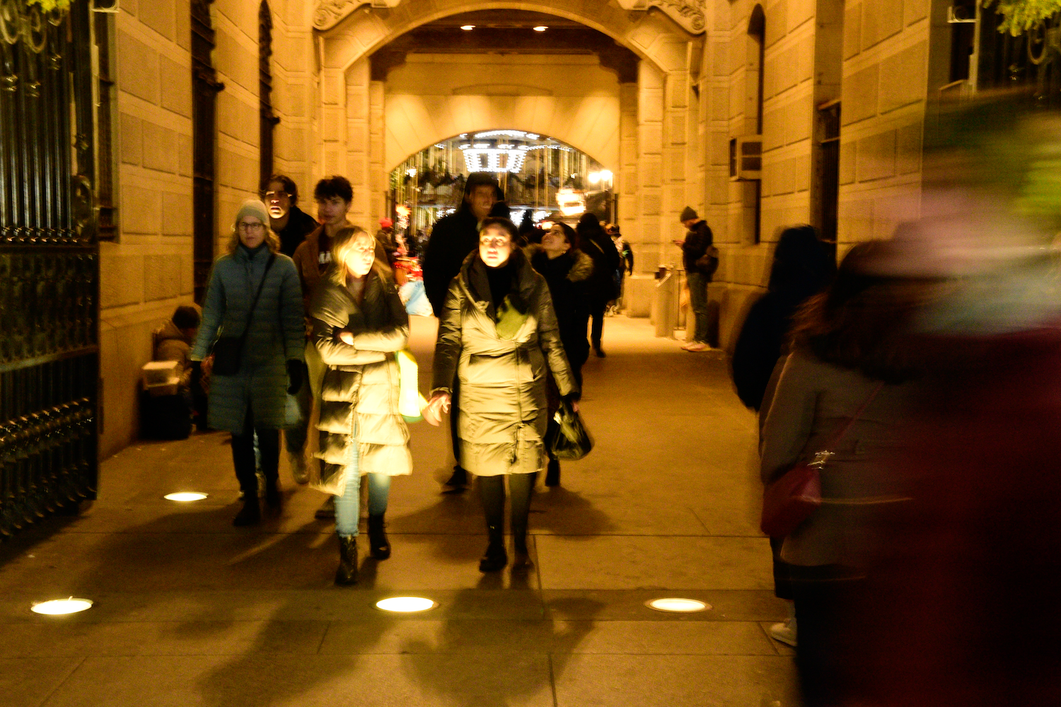 People walking through a stone archway lit by ground lights at night.