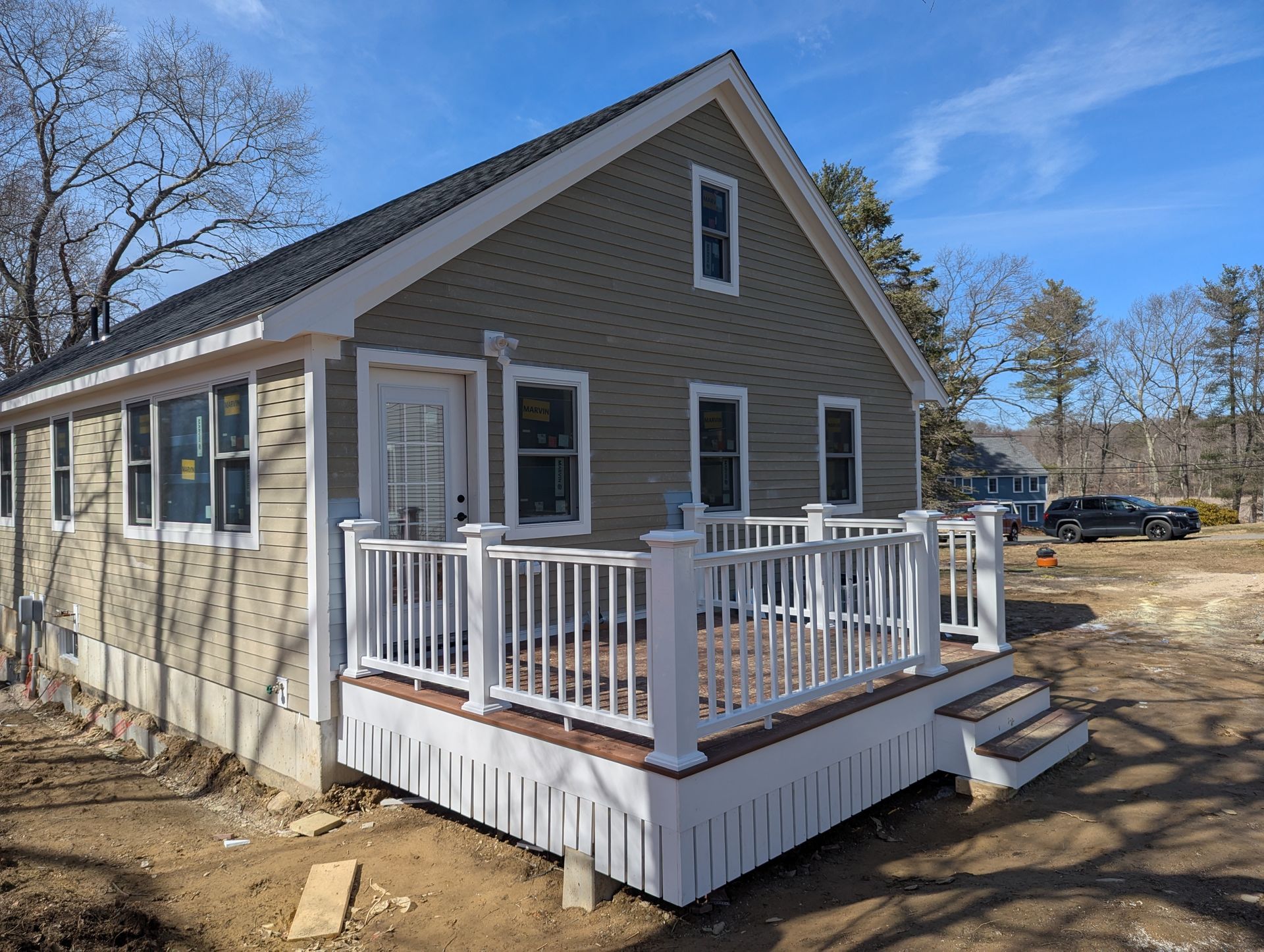 Small beige house with white porch railings on a dirt lot under a blue sky.