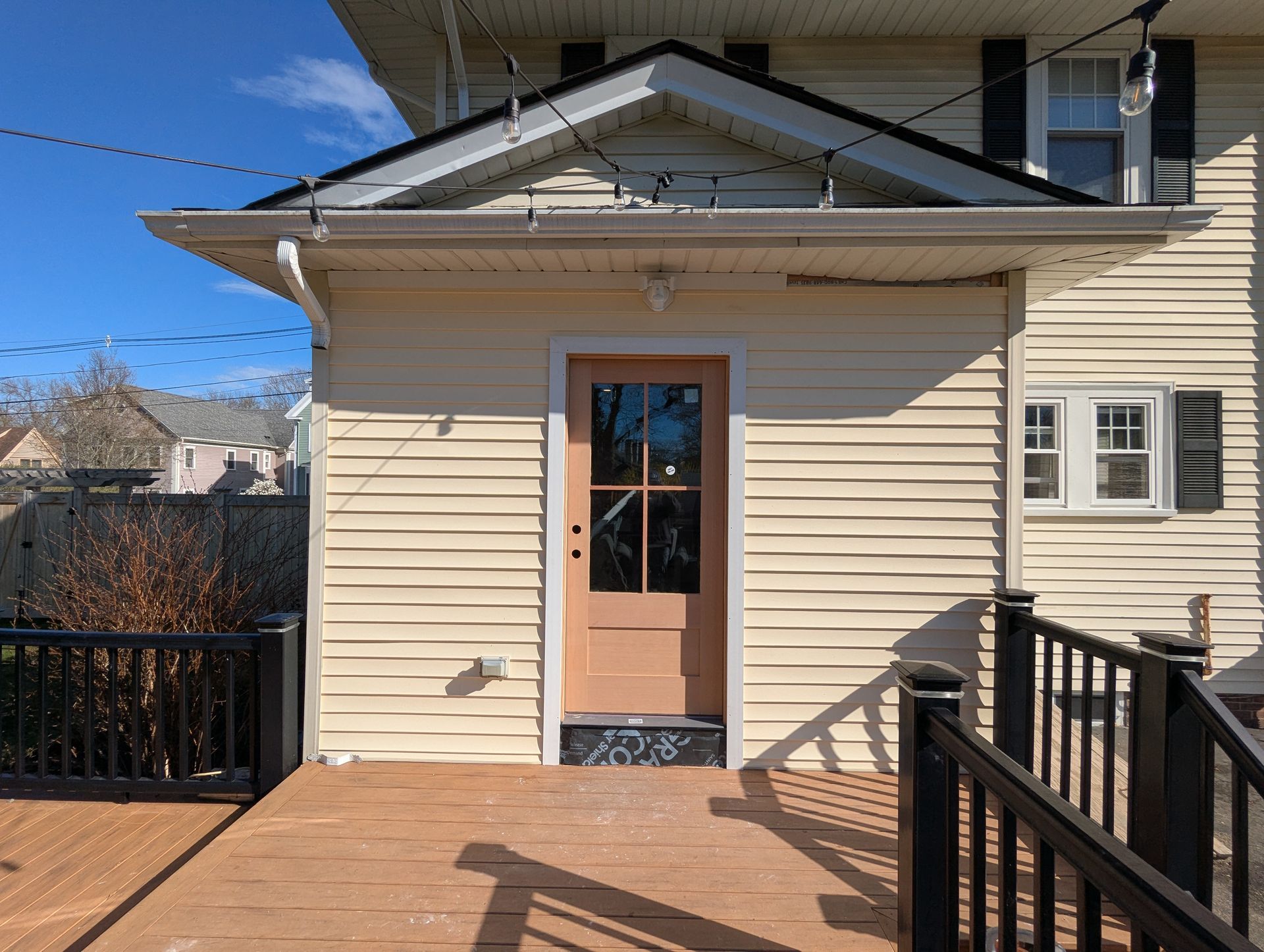 Beige house entrance with wooden door, small porch, and black railing under a bright blue sky
