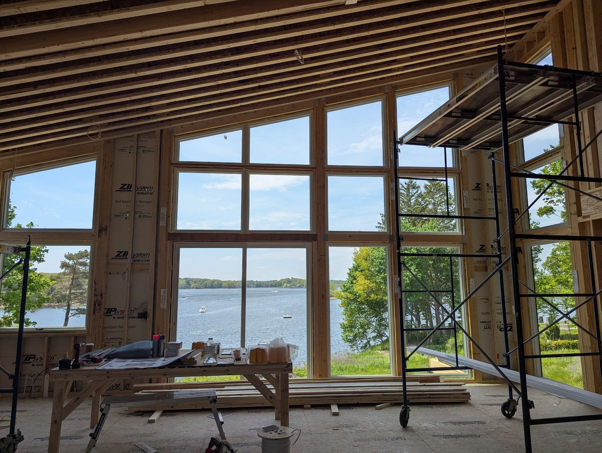 Interior under construction with large window wall overlooking water and a dock, scaffolding on the right.