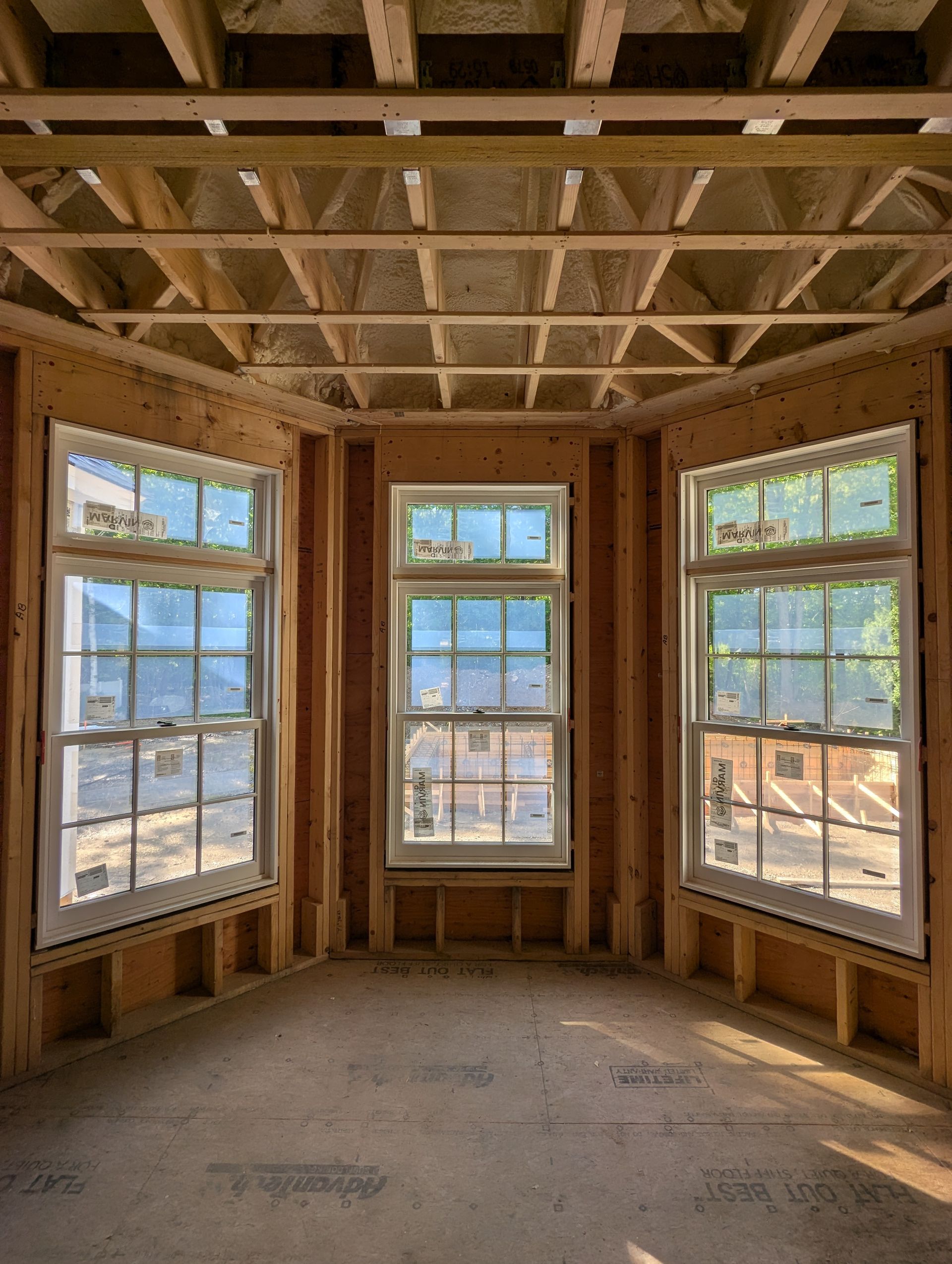 Empty room under construction with exposed ceiling joists and three large windows overlooking a neighborhood.