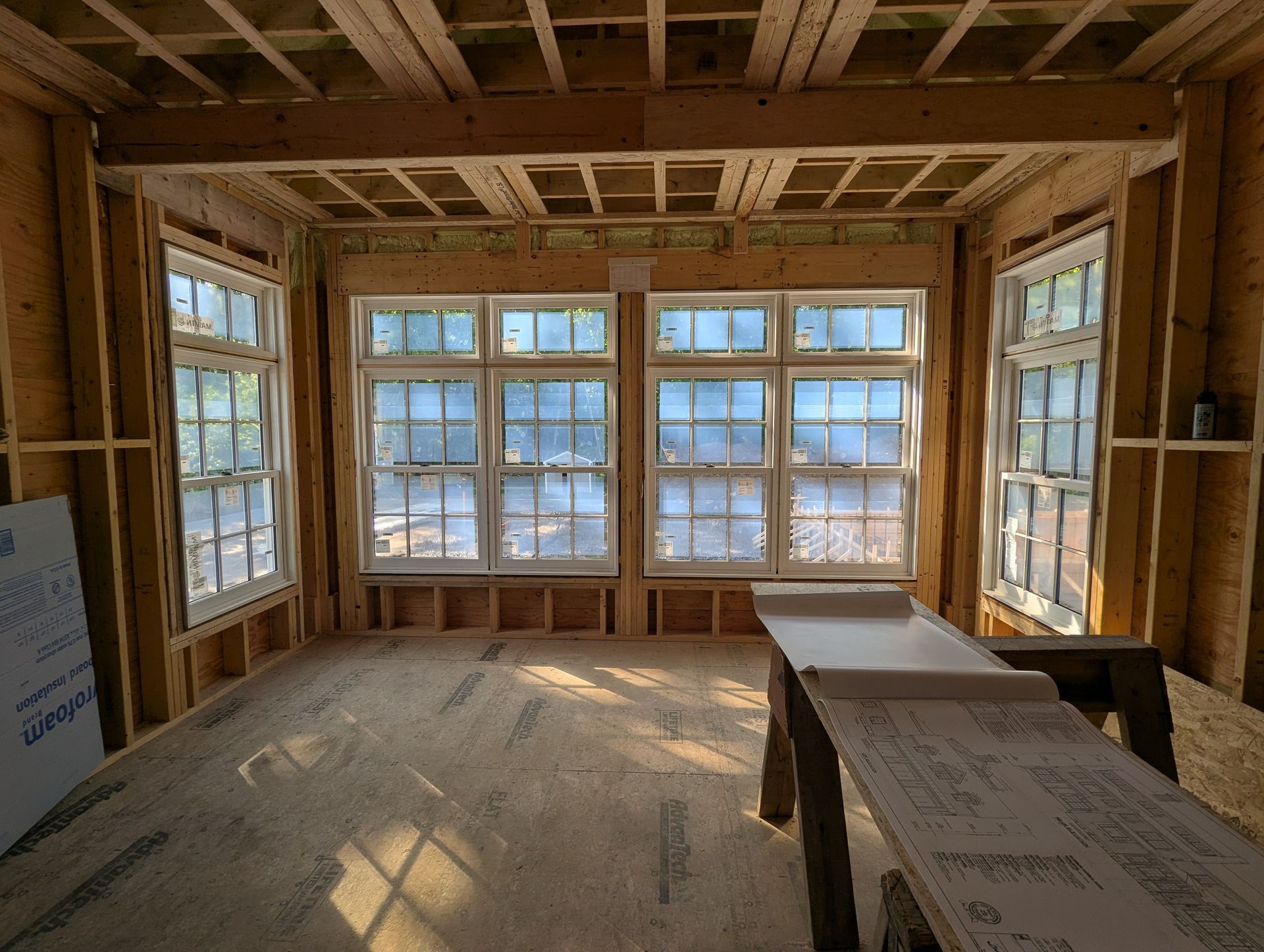 Unfinished room interior with exposed wood framing, large windows, and sunlight on the floor
