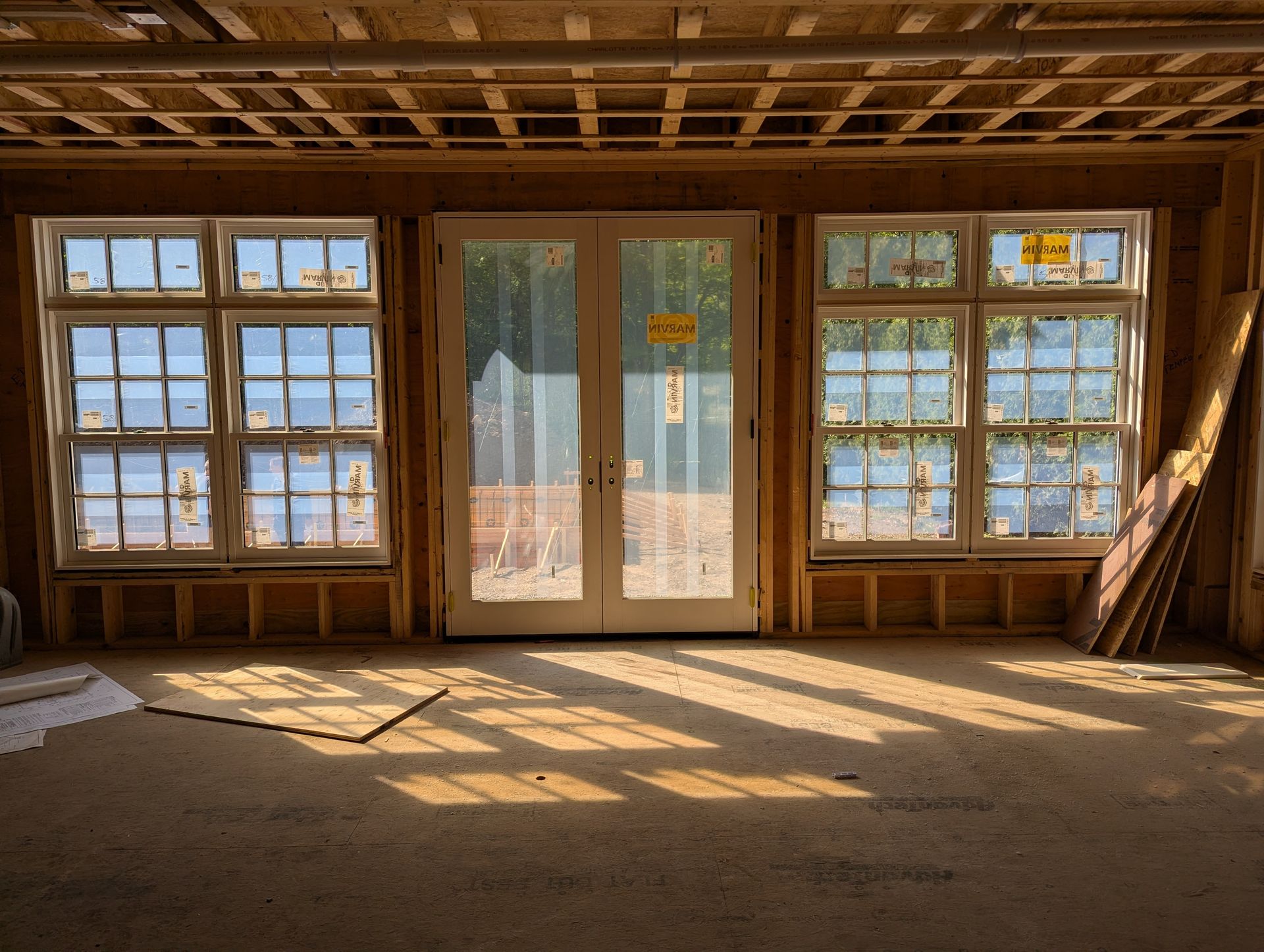 Empty sunlit wooden room with large windows and glass doors, casting patterned shadows on the floor
