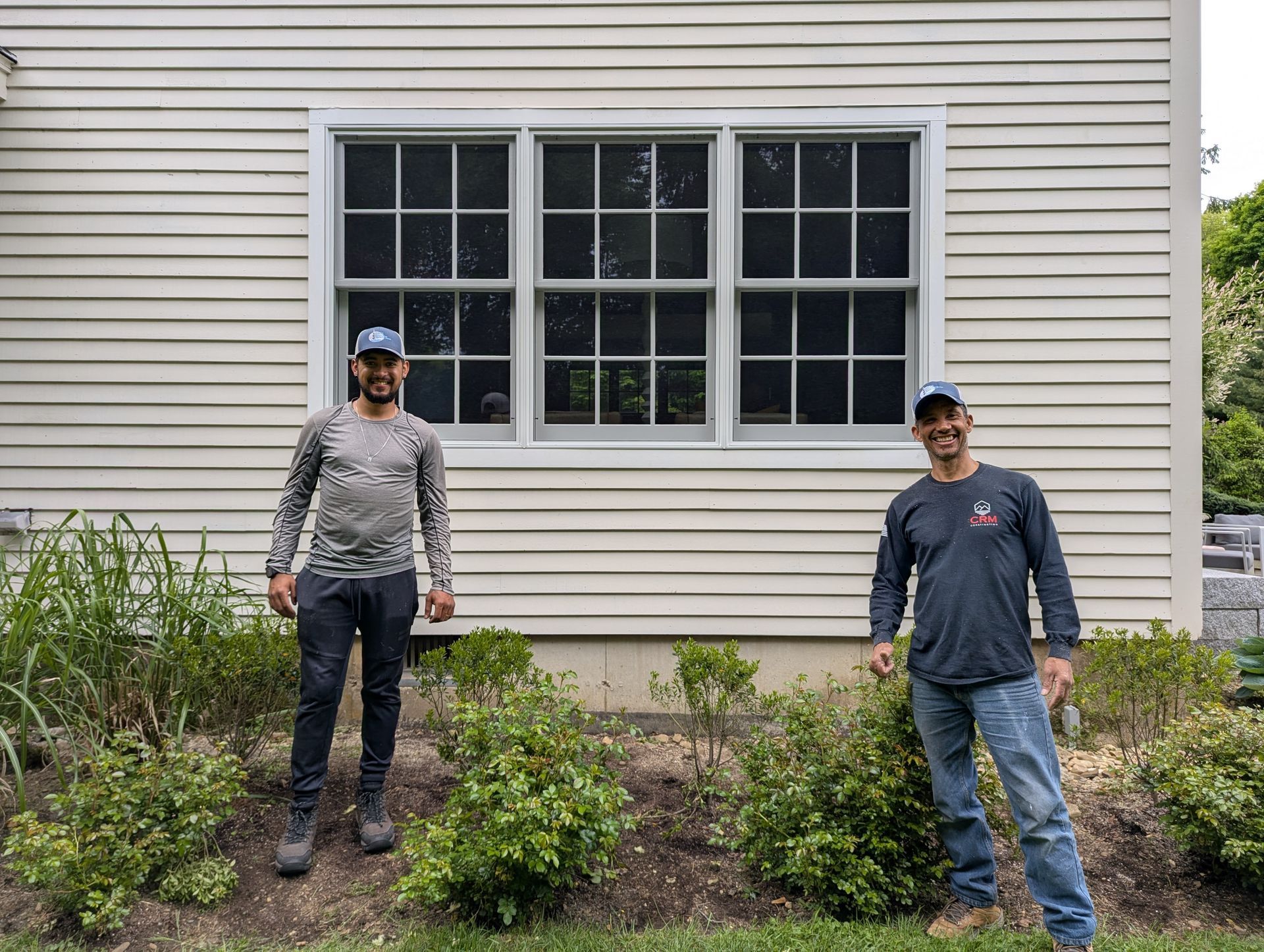 Two people standing in a garden bed beside a house with white siding and a large window.