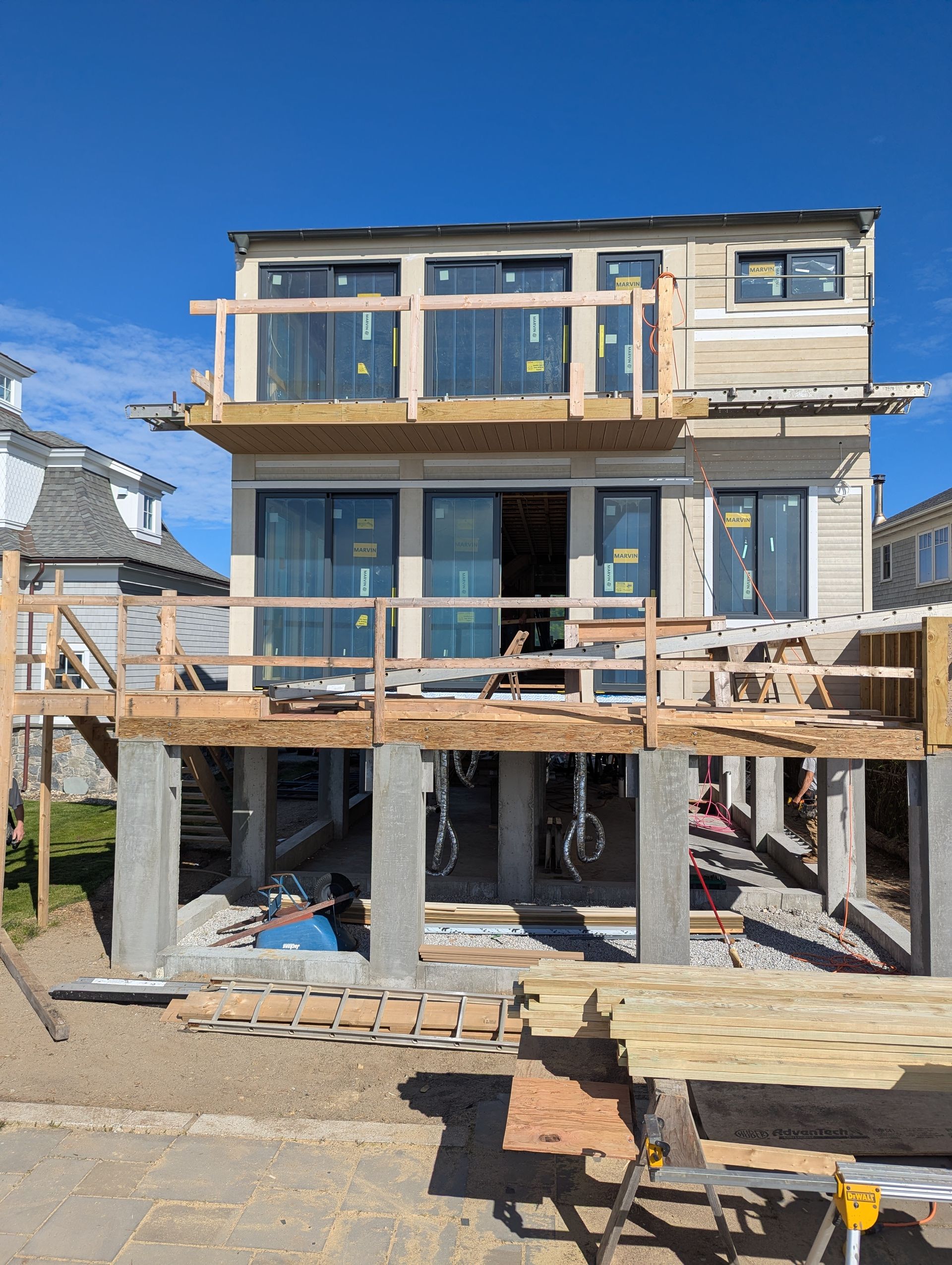 Partially built two-story house with exposed framing, large windows, and a wooden deck under clear blue sky