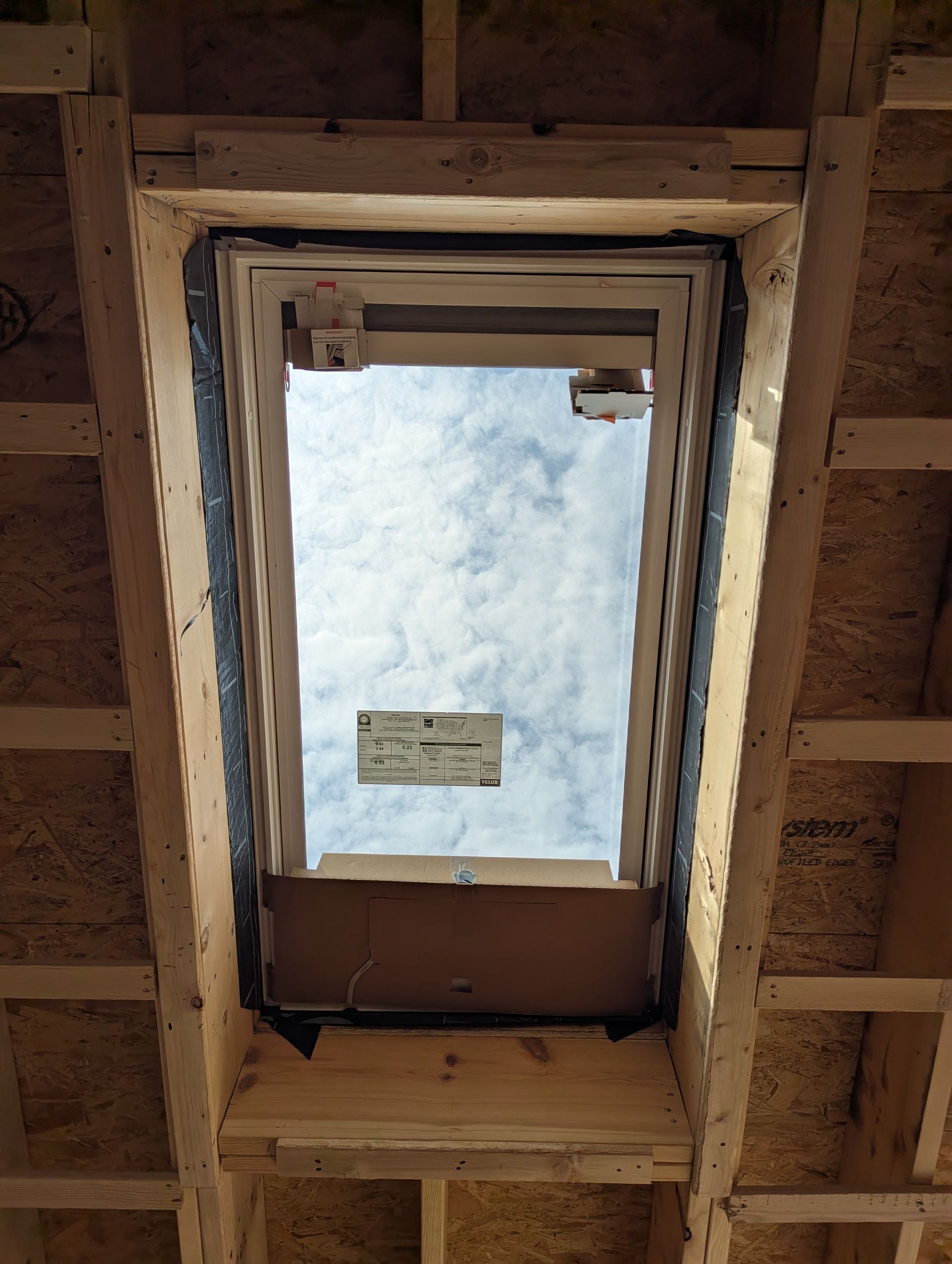 View up through a wooden-framed skylight opening to a cloudy sky, with a small sign visible below.