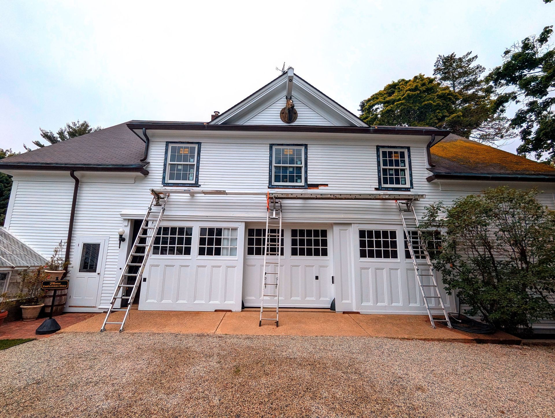 White two-story house with garage doors, dormer windows, and a gravel driveway under a cloudy sky