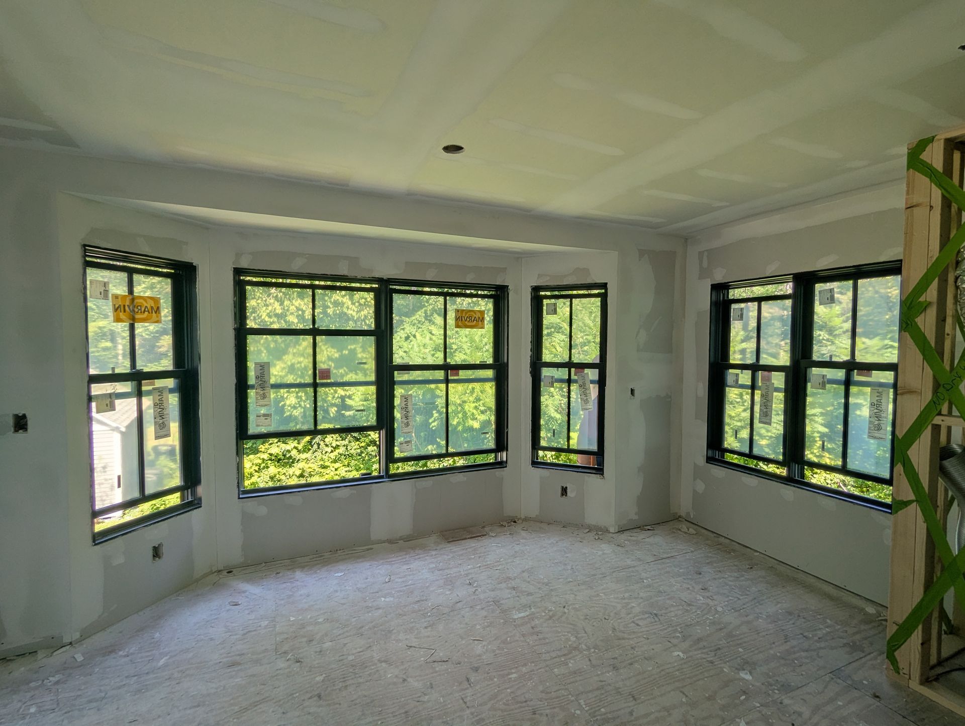 Empty unfinished room with large black-framed windows and white walls, lit by daylight.