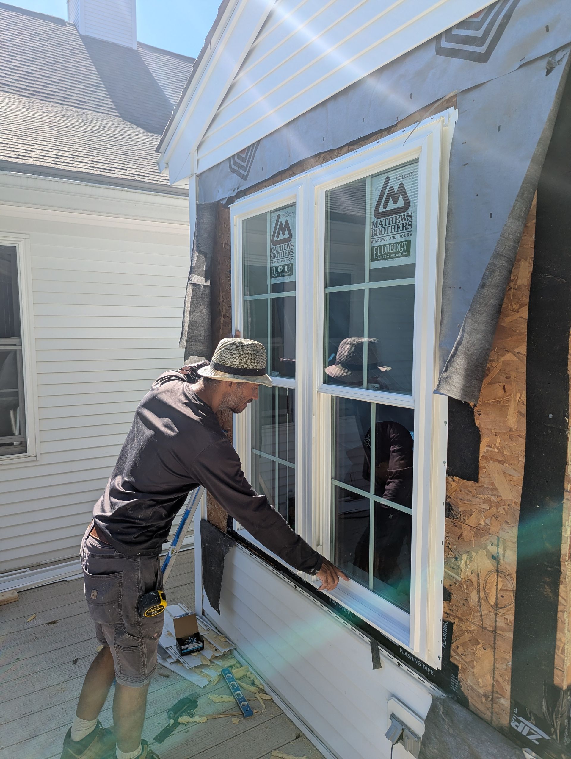 Worker installing a white-framed window on a house exterior while using a tool.