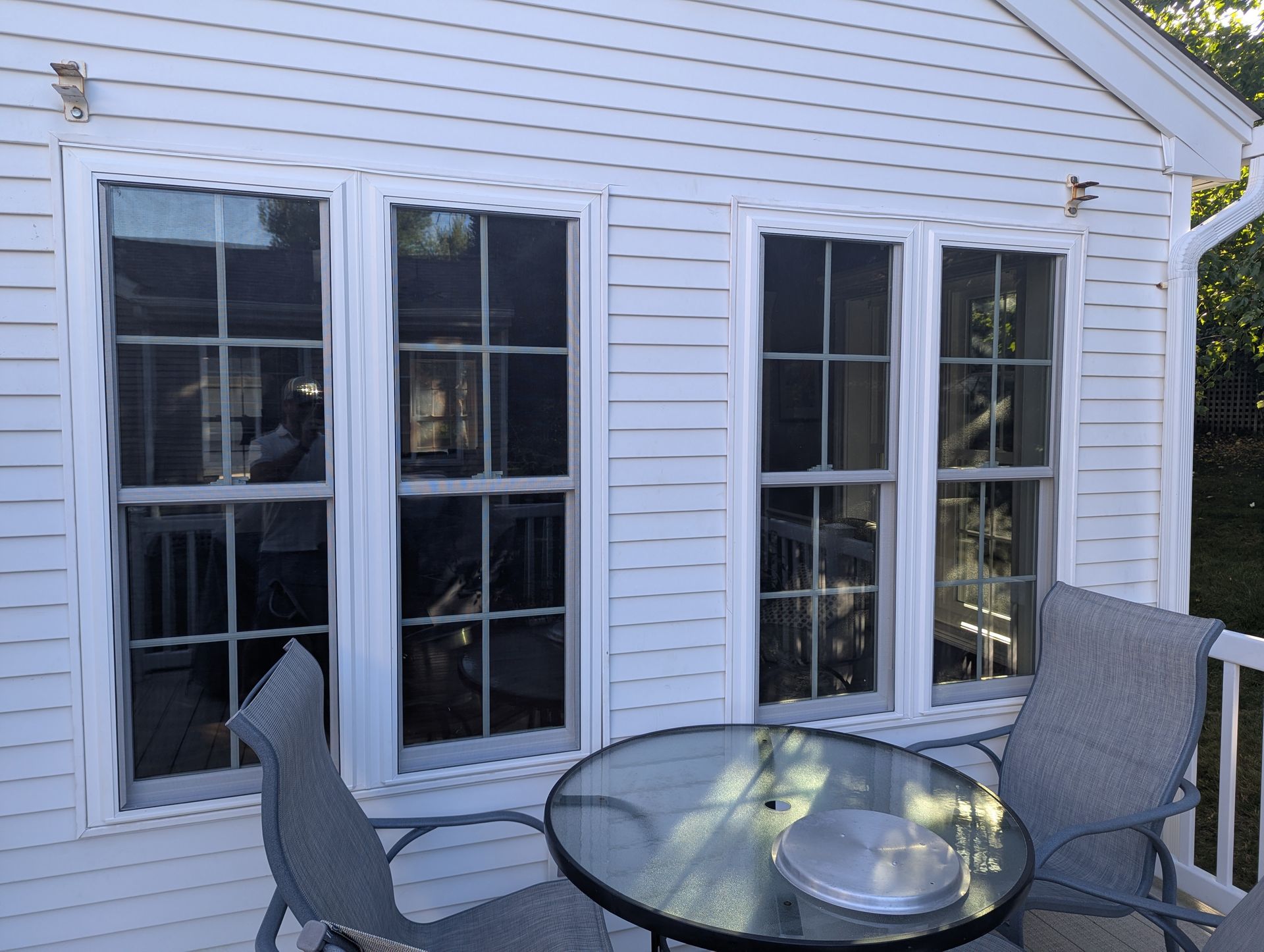 Patio with glass table and chairs beside white house siding and three windows